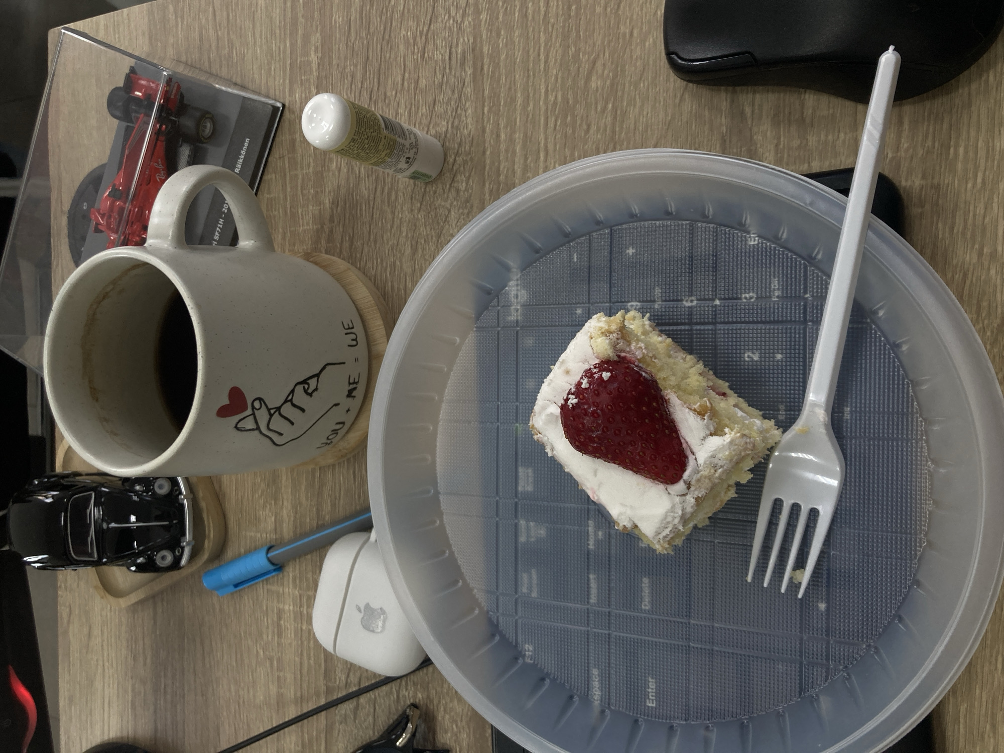 A top-down view depicts a desk in Yerevan, Armenia, with a collection of items. A mug containing a dark beverage sits beside a plastic model car and a pen near wireless earbuds. A clear plastic plate holds a slice of cake topped with a fresh strawberry and a white plastic fork. Adjacent to the plate are a small tube and a plastic container holding a toy car. A computer mouse is also visible on the desk. The desktop surface is wood grain.