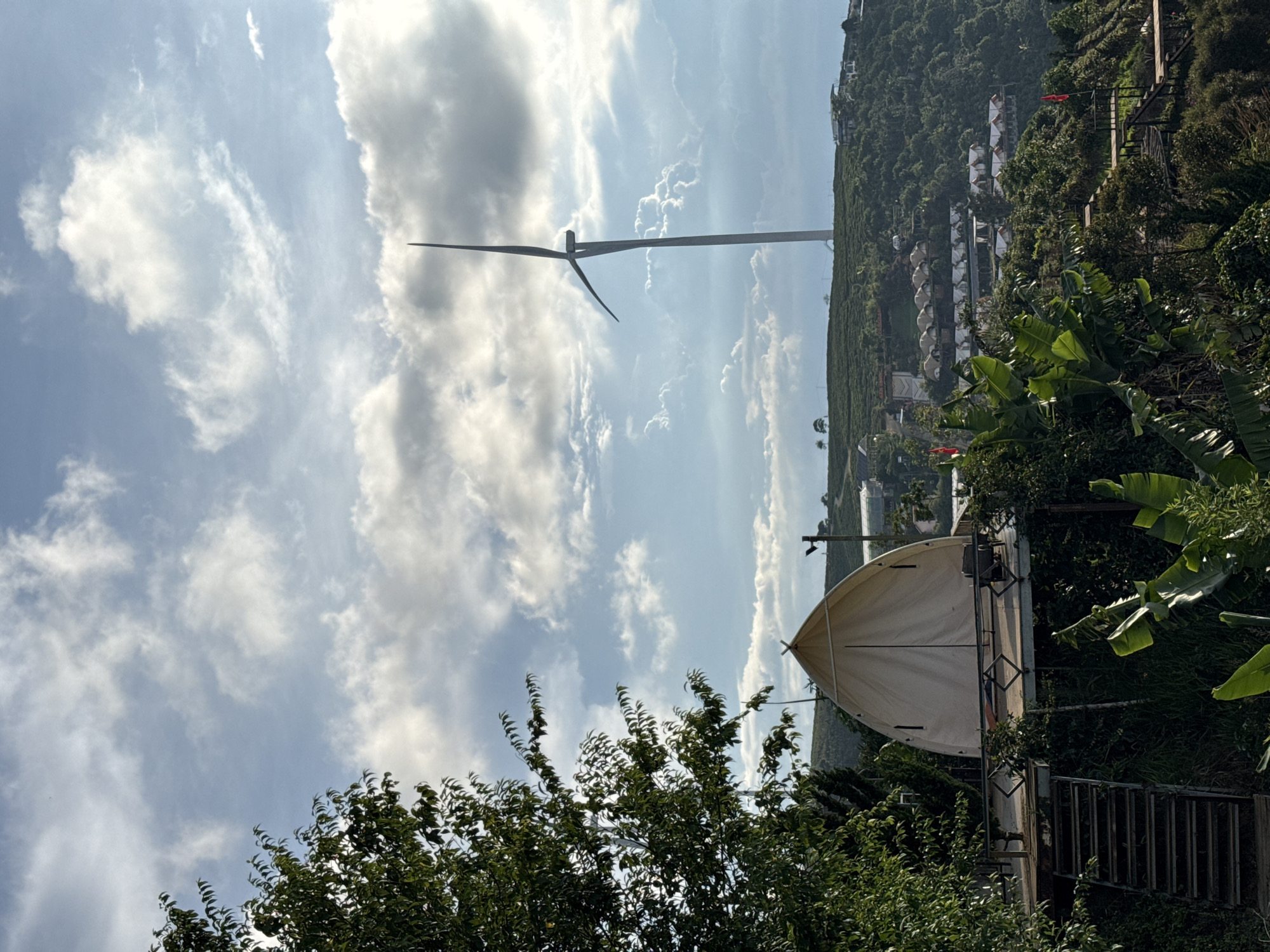 The image depicts a landscape in what appears to be Phát Chi, Vietnam, featuring a large wind turbine against a backdrop of a partly cloudy sky. The turbine stands tall on a hillside densely covered in vegetation. In the foreground, a large tent-like structure is partially visible, surrounded by trees and foliage. A set of stairs leads towards the tent structure. Further up the hillside, there is a cluster of white dome-shaped structures that appear to be buildings or other tent-like accommodations. The overall environment is green and lush.