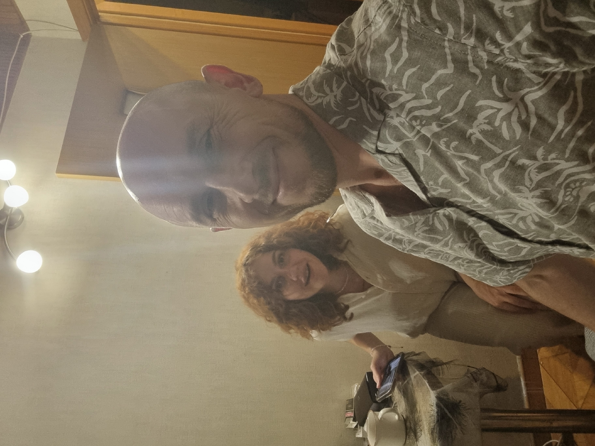 This image captures two individuals indoors, likely within a private residence in Sochi, Russia. A bald man with a short beard and a light-colored short-sleeved shirt patterned with palm trees and abstract shapes is positioned in the foreground, facing and smiling directly at the camera, suggesting a self-portrait. Behind and slightly to his left, a woman with voluminous, curly reddish-brown hair and a light-colored collared shirt is visible, also looking towards the camera with a gentle smile. She holds a dark smartphone in her right hand.
The background features a light, textured wall. Above, a ceiling-mounted light fixture with multiple exposed spherical bulbs illuminates the scene, and a white electrical wire runs along the wall/ceiling line. To the right, a light-colored wooden door frame is partially visible. On the left side of the frame, a round table is covered with a patterned tablecloth. A white ceramic mug with a visible rim, a dark rectangular electronic device, and small stacked items are on the table next to the woman. The floor, visible at the bottom left, appears to be tiled in a light brown or orange hue.