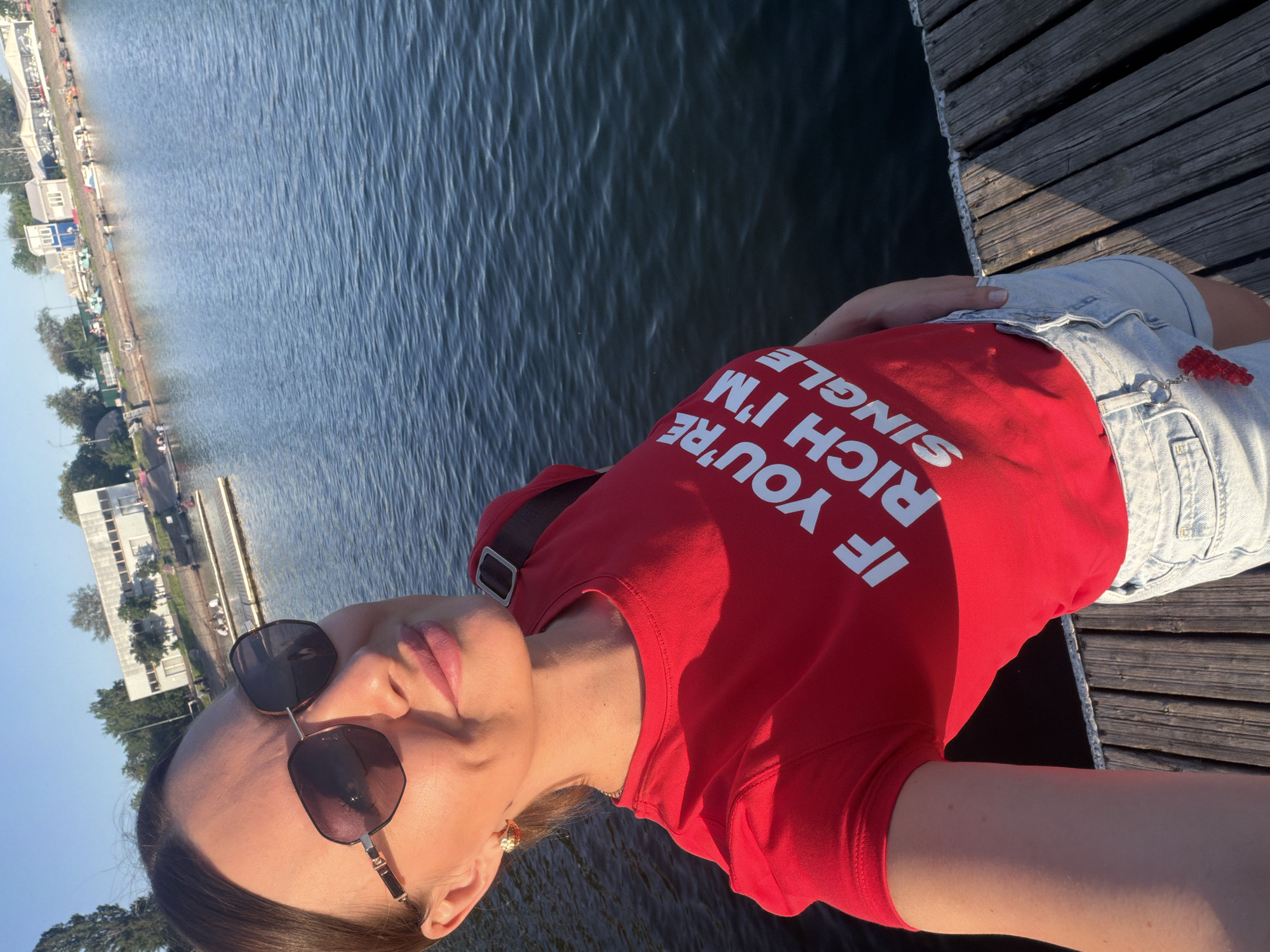 A woman is taking a selfie while on a wooden dock. She is wearing sunglasses, a red shirt with the text "IF YOU'RE RICH I'M SINGLE", and denim shorts. The background shows water with sunlight reflection, some buildings, and vegetation. The location is most likely near the Don River in Rostov-on-Don, Russia.