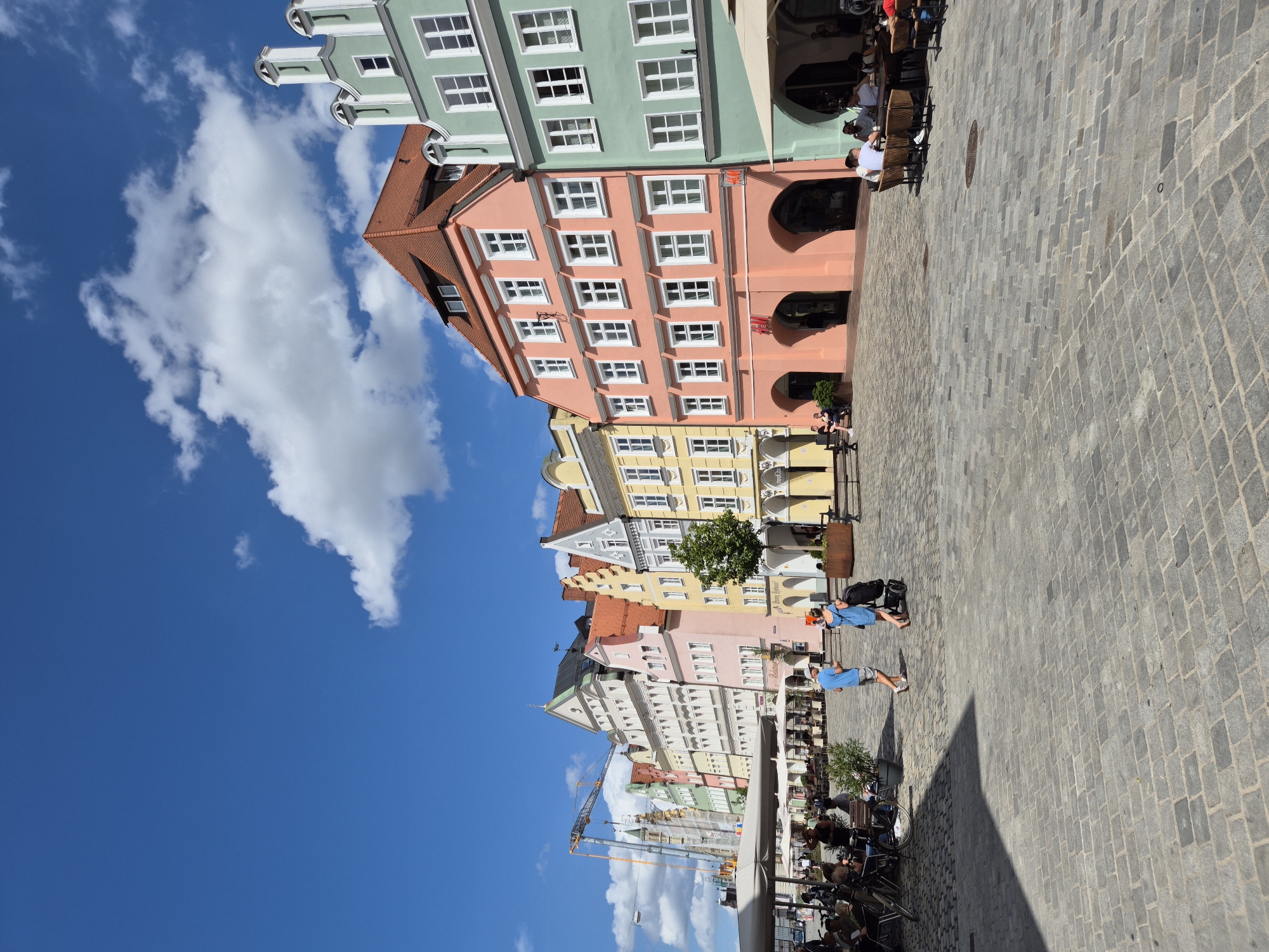 This outdoor, eye-level, wide-angle shot depicts the Altstadt (Old Town) in Landshut, Germany, on a clear, sunny day. The scene is dominated by a row of distinctively gabled, multi-story buildings, painted in various pastel colors, lining a cobblestone square.
From right to left, the buildings display a range of hues: a light green building with ornate white stepped gables and multiple windows; a prominent salmon-pink building with a red-tiled gable roof, numerous white-framed windows, and three arched entrances on the ground floor, one bearing a red "H&M" logo; and a yellow building with similar architecture. Further down the street, additional buildings are visible in shades of white, light green, pink, and grey, each with distinct gabled roofs and numerous windows. Towards the far left, a tall yellow construction crane rises against the sky, indicating ongoing development.
The ground is paved with grey cobblestones, arranged in a regular pattern. Shadows are cast by unseen objects or structures on the left side of the pavement.
Several people are present. In the mid-ground, two individuals walk away from the camera: one person in a blue shirt and shorts, and another in a blue dress pushing a dark stroller. On both the right and left sides of the square, groups of people are seated at outdoor tables with brown chairs, some under large beige or white umbrellas, seemingly engaged in dining or socializing. A black bicycle is parked near tables on the left. Small green plants are visible in dark planters.
The sky is a bright, clear blue with scattered white cumulus clouds. Bright sunlight illuminates the scene, creating distinct shadows.