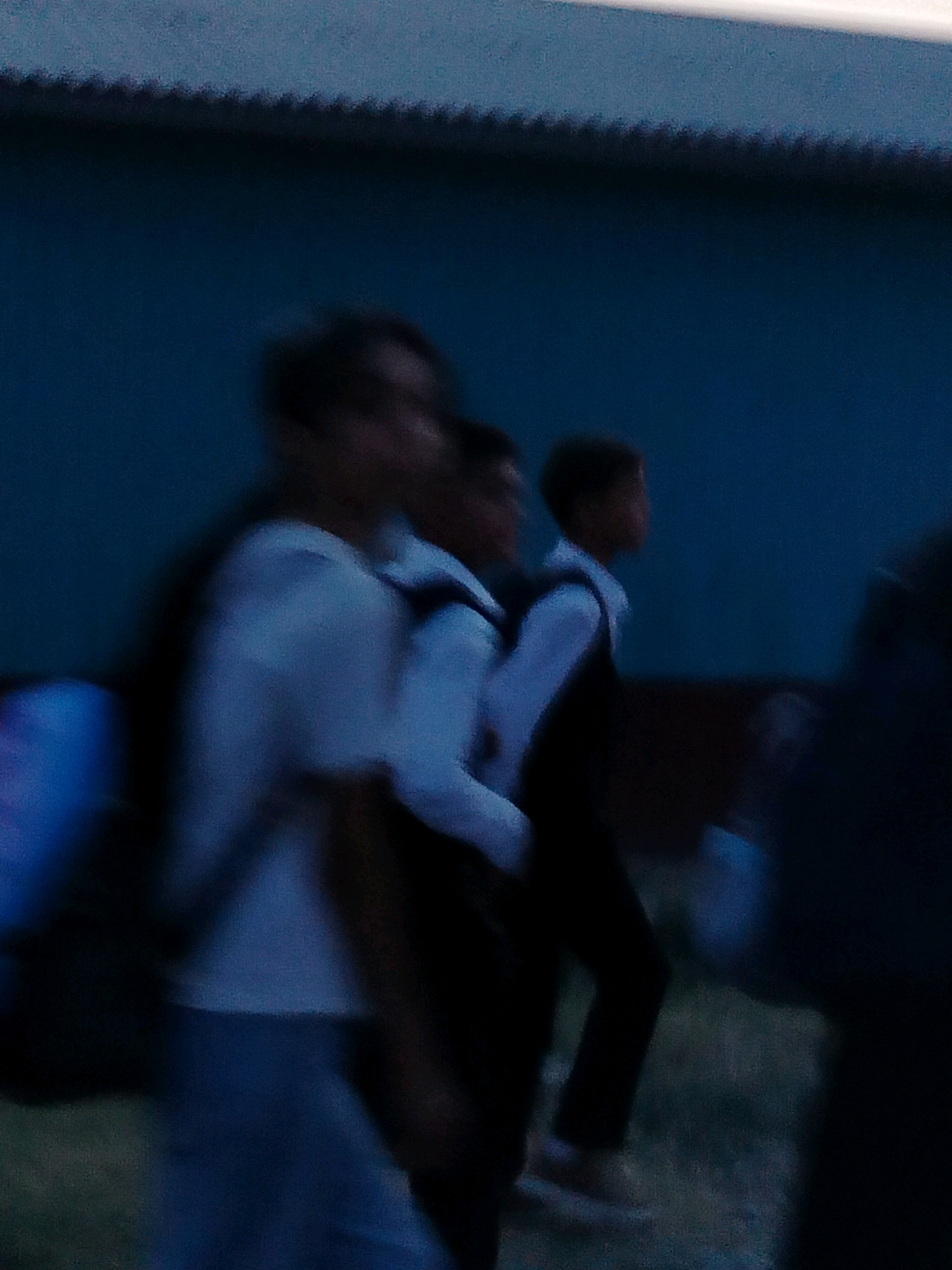 The image depicts a blurred scene with several individuals, likely students, walking outdoors. The background consists of a dark blue wall with a textured, ridged top edge. The lighting suggests it is dusk or early evening. In the foreground, a person wearing a white shirt and light-colored pants is visible, carrying a backpack on their left shoulder. To their right, another individual in a dark vest and white shirt is also walking. Further to the right, parts of other figures are visible, also appearing to be in motion. The ground is a mixture of dark soil and some grassy patches. The overall impression is of a group of people moving away from the camera in an urban or semi-urban environment, possibly near a school. No specific landmarks for Көлкент, Kazakhstan are identifiable.