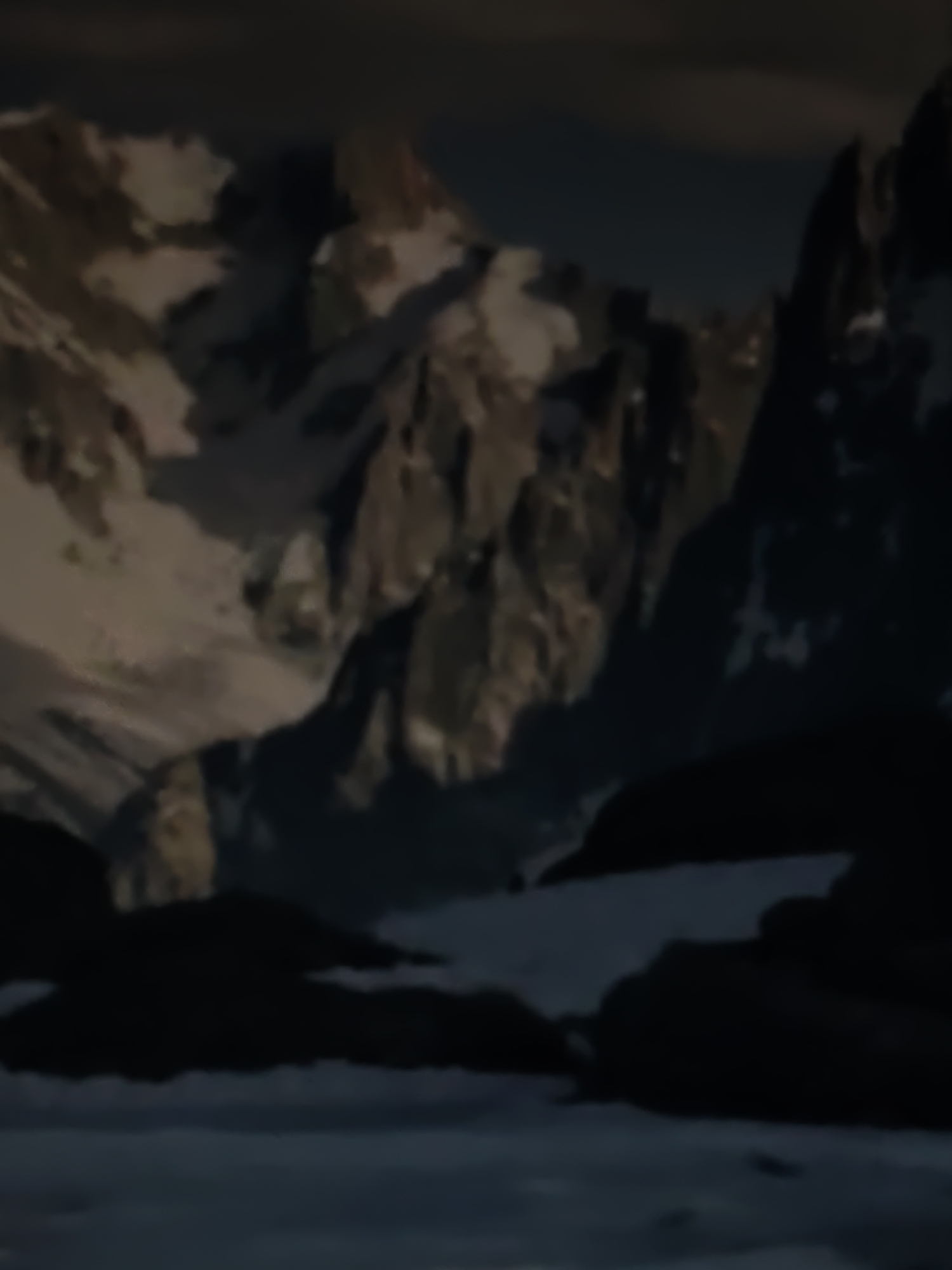 The image shows a blurred, dark, and moody mountainous landscape. Jagged peaks, some covered in snow, rise in the background under a dusky sky. The foreground is occupied by dark, rocky terrain, also partially covered in snow. The overall impression is one of a harsh, remote, and possibly cold environment. There are no people or man-made structures visible in the image. The location in the city of Kurganinsk, Russia is not discernible from the visual information provided in the image.