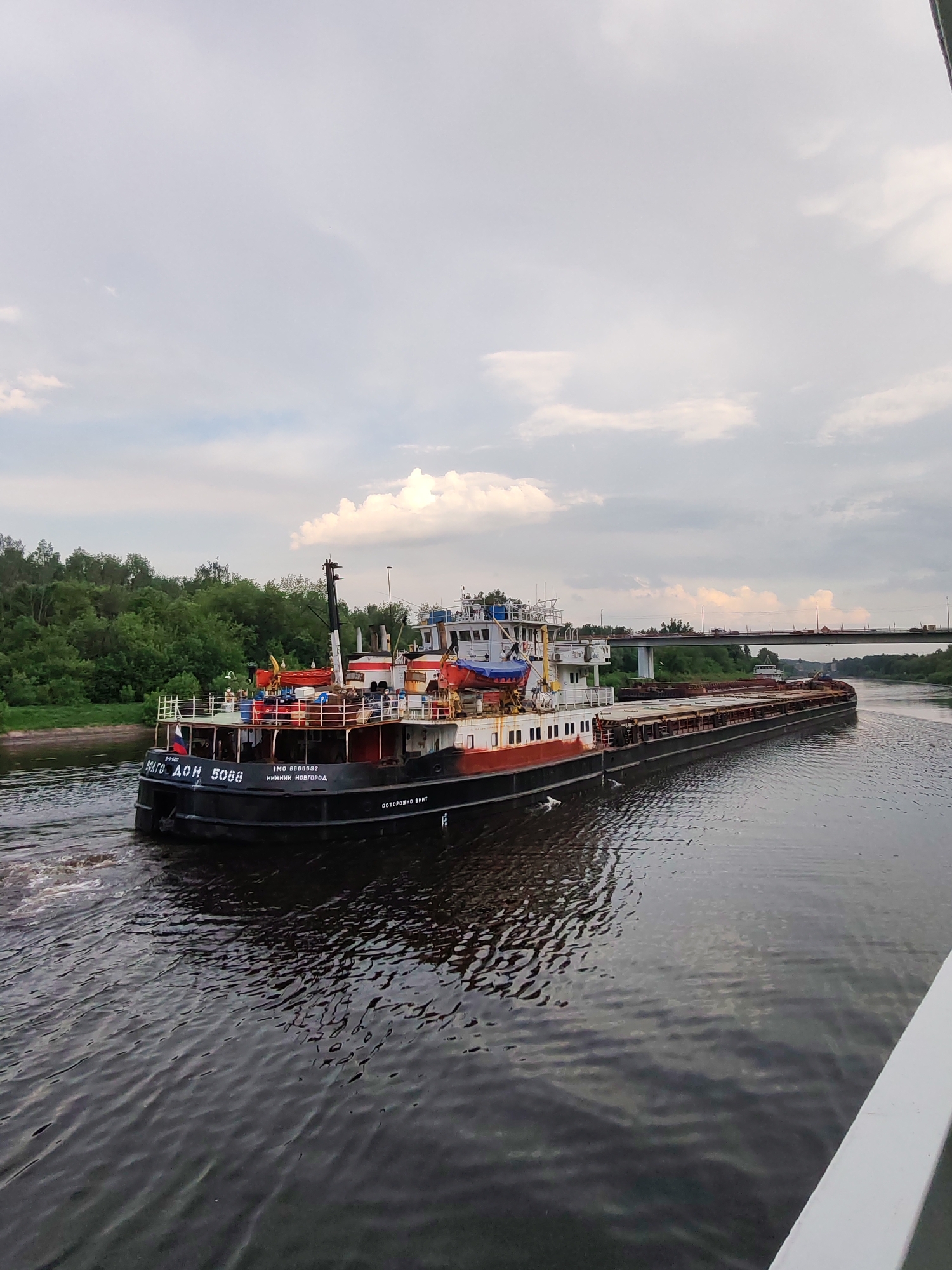 A barge, with a tugboat connected at its stern, navigates a river in the city of Yakhroma, Russia. The tugboat, marked "Волго-Дон 5088" and "Нижний Новгород," displays the Russian flag. A bridge carrying road traffic is visible in the background. The river is bordered by green trees and vegetation. The sky is overcast with clouds. The photograph is taken from an elevated position, potentially from another vessel or a bridge.