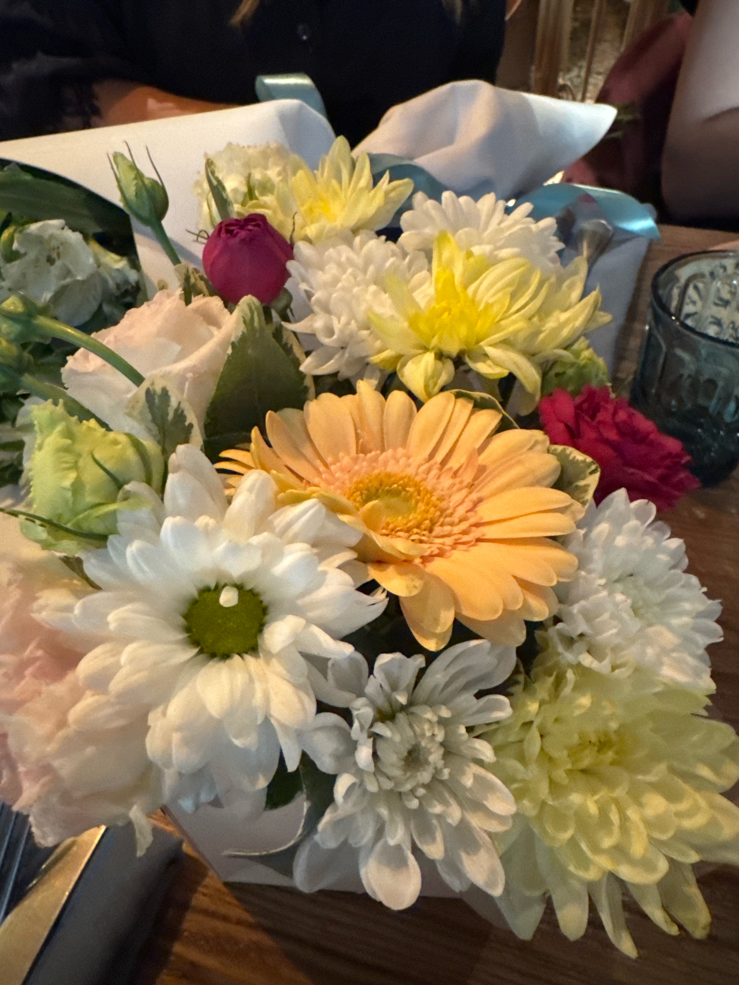 A close-up, eye-level shot captures a vibrant bouquet of assorted flowers. The arrangement includes a large orange gerbera daisy, surrounded by white chrysanthemums, yellow mums, and pink roses. A dark pink rose bud is visible near the top left, alongside a green rose bud. The flowers are presented in a white vase, which is partially obscured by the bouquet. The background is softly blurred, showing hints of people and a blue glass. The overall lighting is dim, suggesting an indoor setting, possibly a restaurant or home, and there is no discernible indication of the specific location in Ufa, Russia.