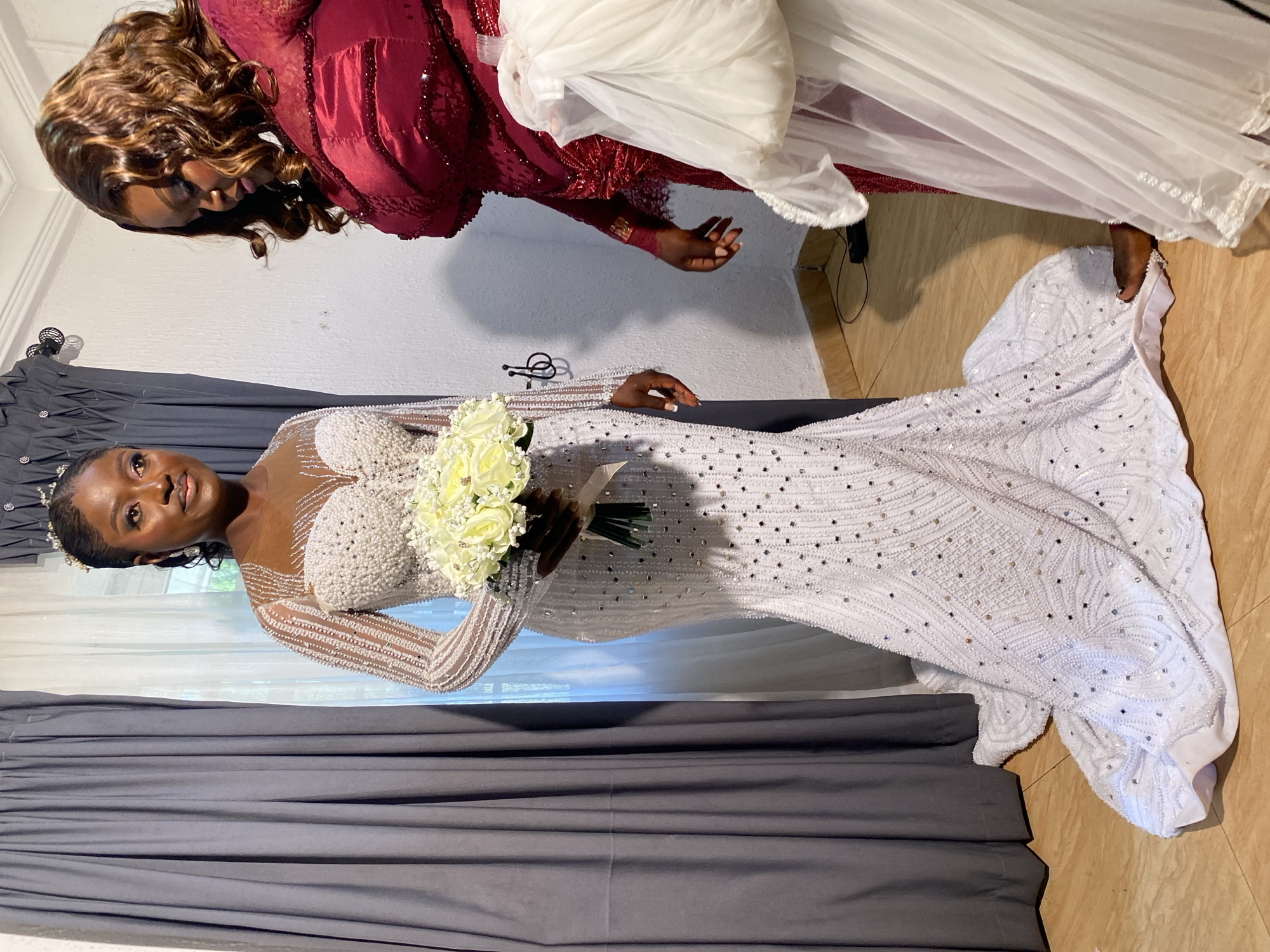 Two women are standing indoors, likely in a home in Bukuru, Nigeria, preparing for a wedding. The woman on the left, the bride, is wearing a white, floor-length wedding gown heavily embellished with pearls and sequins. The gown features a sweetheart neckline and long, sheer sleeves adorned with the same embellishments. She is holding a bouquet of white roses and is wearing a tiara and earrings. The woman on the right is wearing a dark red, long-sleeved dress also with embellishments. She is holding a piece of white fabric, possibly part of the bride's attire. The background consists of gray curtains on the left and a white wall on the right. The floor is tiled. The lighting suggests it is daytime, possibly with natural light coming from a window behind the curtains.