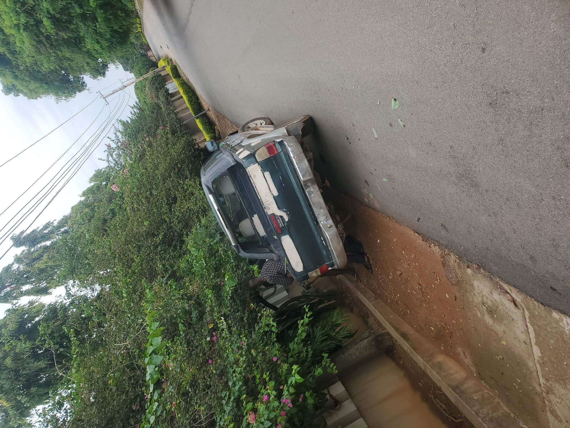 The image depicts a scene on a roadside in Jos, Nigeria. A dark green pickup truck, appearing weathered and possibly damaged, is parked on the side of a paved road. The truck has a dented rear bumper and its paint is chipped in several places. A person wearing a checkered shirt and dark pants is standing next to the rear of the truck, leaning towards it. The environment is characterized by lush green trees and bushes lining the roadside, with some pink flowers visible among the foliage. A concrete wall runs along the left side of the scene, bordering a dirt and gravel area. Power lines are visible overhead, and a utility pole stands further down the road. The sky is overcast.