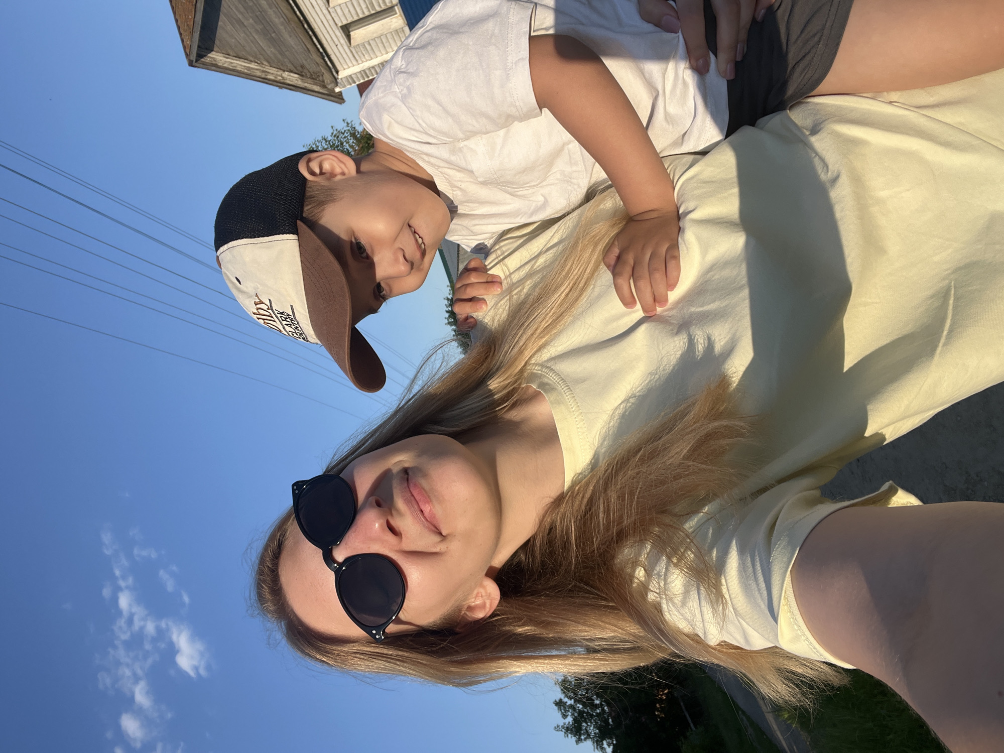 A woman and a young child are captured in a low-angle, selfie-style photograph against a bright blue sky with scattered clouds. The woman, positioned in the foreground and on the left side of the frame, has blonde hair and is wearing large, black, round sunglasses and a light yellow t-shirt. She is smiling and looking slightly upwards. The child, on the right side of the frame, is being held by the woman and is wearing a white t-shirt and a beige and brown baseball cap. The child is looking to the left of the frame and smiling. In the background, there are visible power lines, a glimpse of a wooden house, and green vegetation. The lighting suggests it is late afternoon or early evening, with warm sunlight casting shadows. The location is not specified as Бруснятское, Russia, but is described as an outdoor, possibly suburban or rural setting.
