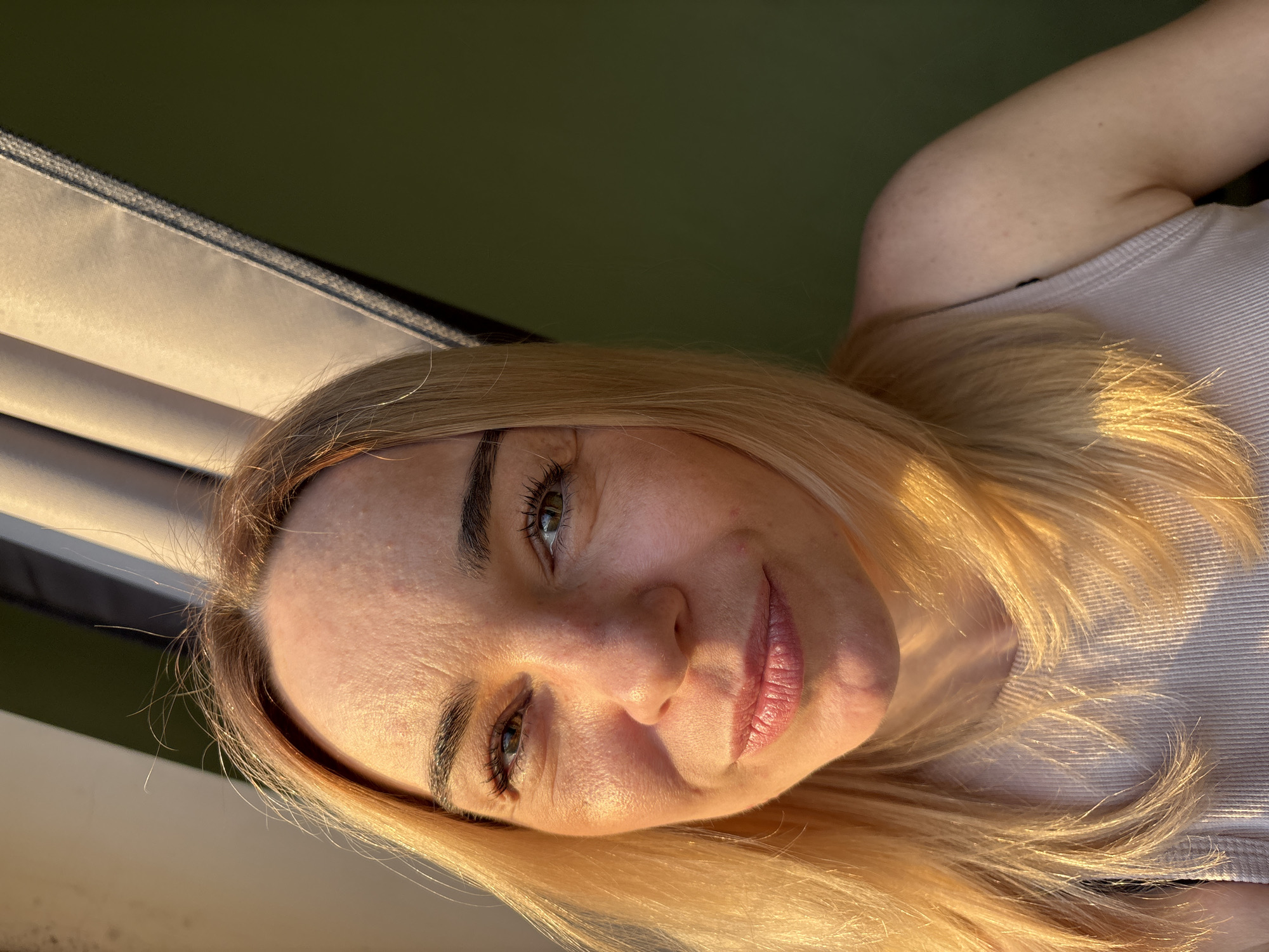 This close-up portrait captures a woman with blonde hair and fair skin, looking directly at the viewer with a soft, gentle smile. Her head is tilted significantly to the bottom right of the frame. She has dark eyebrows and green or hazel eyes. Warm, golden light, indicative of the "golden hour" (early morning or late afternoon), illuminates her face and hair, creating highlights and shadows that emphasize her features. She appears to be wearing a light-colored, possibly beige or light pink, ribbed tank top or camisole, visible over her right shoulder.
The background on the top right consists of a dark, muted green, which could be shaded foliage or a distant natural element. To the top left and extending down the left side, a light-colored, woven or canvas-like fabric structure is visible, likely part of an outdoor seating arrangement such as a hammock or recliner, also touched by the golden light. The overall scene conveys a relaxed and pleasant atmosphere. No specific activity is taking place beyond the woman posing for the photograph. There is no visible text. The setting appears to be outdoors, with strong natural light, but the exact location is not discernible.