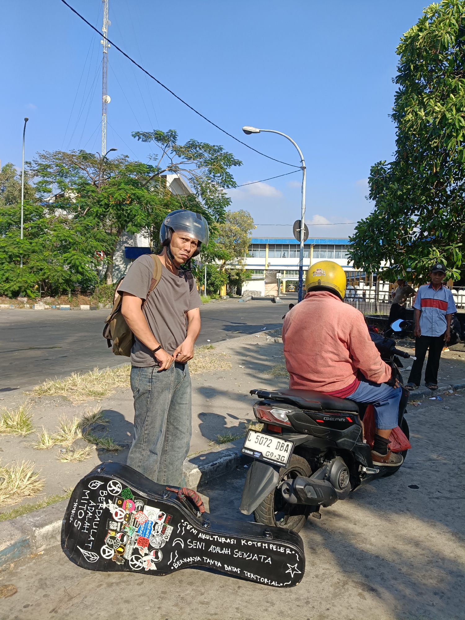 A man wearing a grey t-shirt, blue jeans, and a black helmet stands on a paved surface, looking towards the camera with a neutral expression. He has a backpack slung over his left shoulder. To his right, a black motorcycle is parked, with another person wearing a coral-colored jacket and a yellow helmet seated on it, facing away from the camera. A third man, wearing a light blue and red striped shirt and dark pants, stands to the right of the motorcycle, looking towards the right side of the frame. In the foreground, a black guitar case rests on the ground, adorned with numerous stickers and handwritten text. Behind the people, a street is visible, bordered by trees and buildings. A tall communications tower is prominent in the background. The scene appears to be an outdoor urban environment, possibly in Kedungrejo, Indonesia, given the Indonesian license plate on the motorcycle which reads "L 5072 DBA". The sky is clear and blue, suggesting daytime.