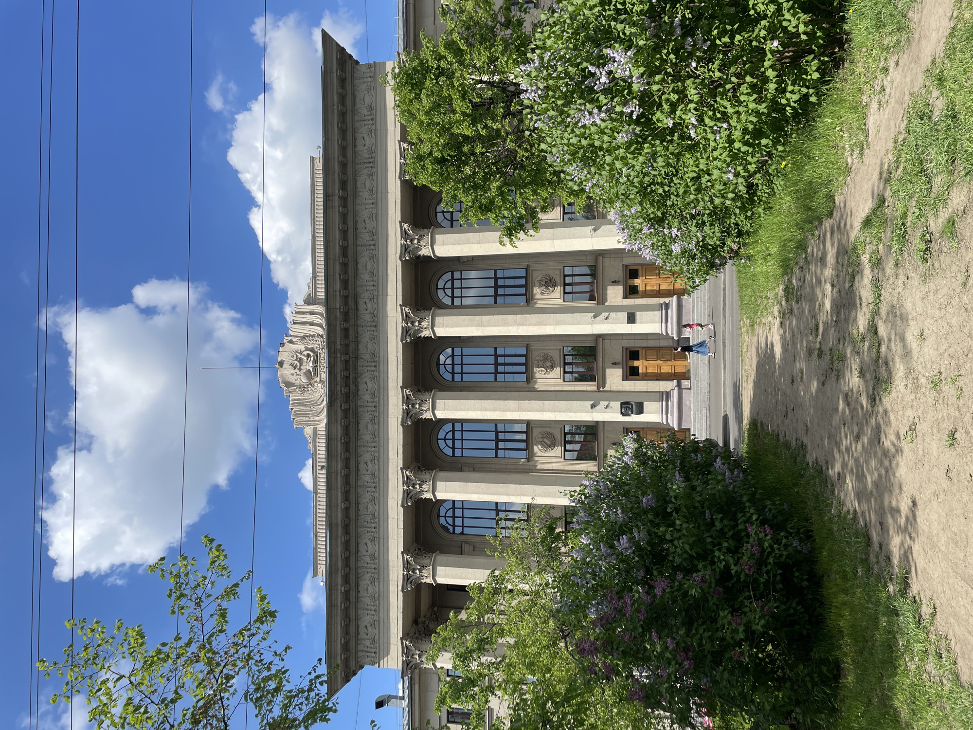 The image depicts the Khimki Administration Building in Khimki, Russia. The building is a neoclassical structure with tall columns and arched windows. It has a decorative cornice and a stepped facade. In front of the building are several green trees and bushes with purple flowers. There are two people walking towards the building entrance. Above the building, a blue sky is visible with scattered white clouds. Utility lines are also visible against the sky. A dirt path is visible on the right side of the image.