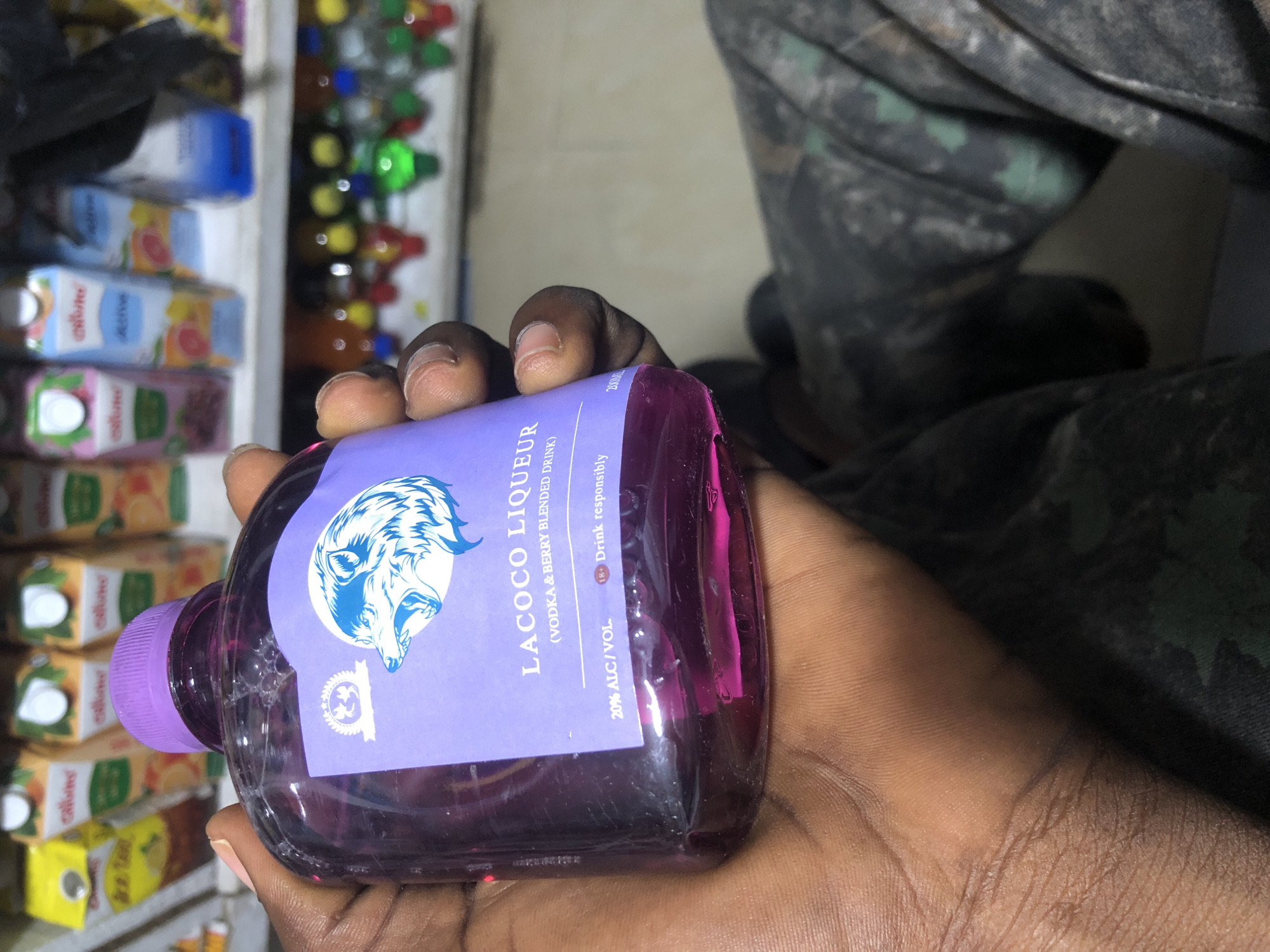 A hand, likely belonging to a person of color, is holding a clear plastic bottle filled with a vibrant purple liquid. The bottle has a purple cap and a label with a blue wolf logo and text that reads "LACOCO LIQUEUR (VODKA & BERRY BLENDED DRINK)". Below this, smaller text indicates "20% ALC./VOL." and "18+ Drink responsibly". The background is blurred but suggests a retail environment, possibly a convenience store or liquor store, with shelves stocked with various beverages, including juice cartons and bottles. The person is wearing camouflage-patterned clothing. This scene is not set in Ado, Nigeria; there are no identifying features of that city present.