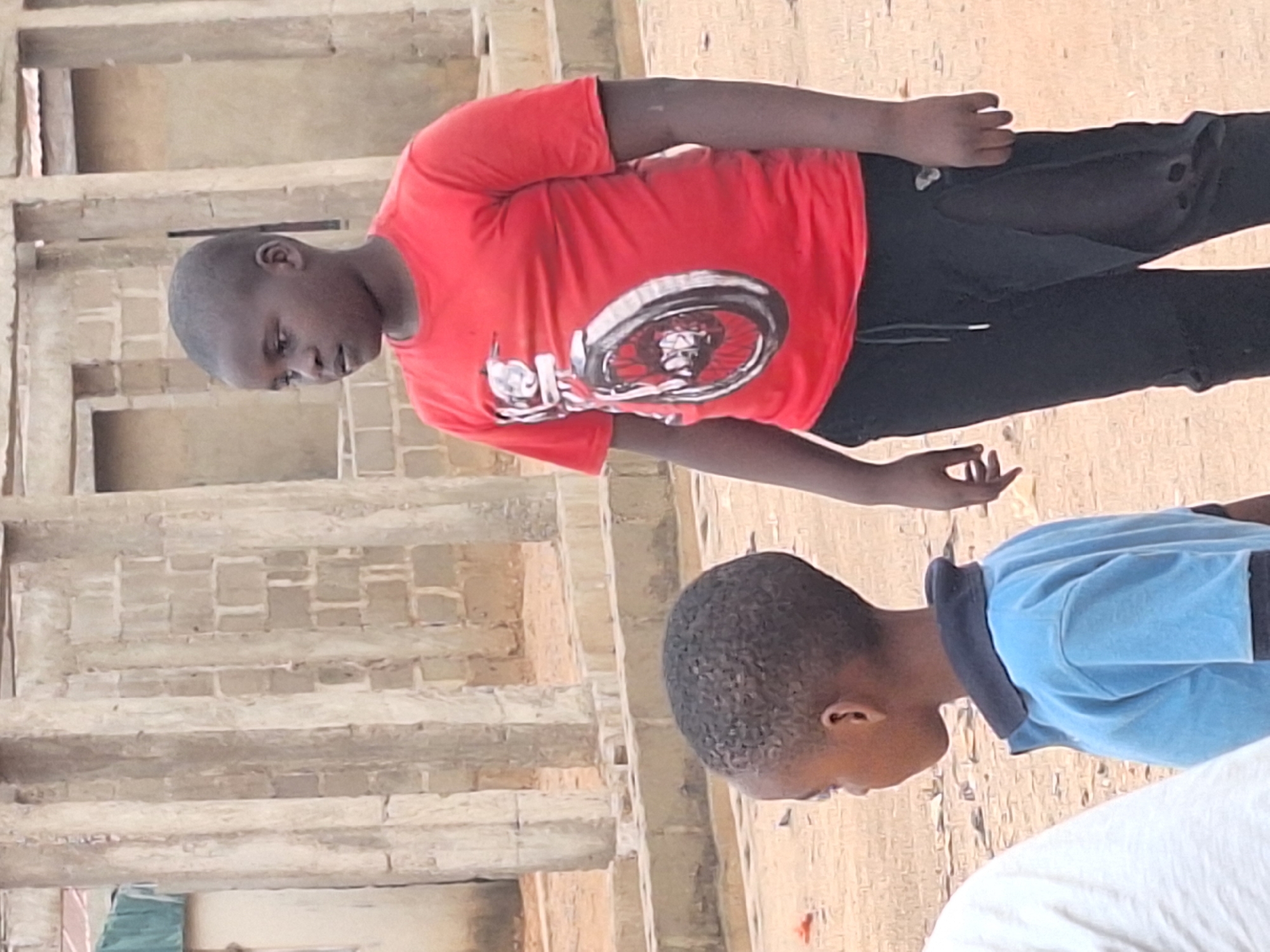 In Sabon Tasha, Nigeria, two young boys are outdoors on what appears to be a dirt or sand-covered area near a building under construction. The building is made of concrete blocks with exposed rebar. One boy is wearing a red T-shirt with a graphic and black pants; he is standing. The other boy is wearing a light blue shirt with a dark blue collar, and he is also standing. The interaction and activities between them are indiscernible.