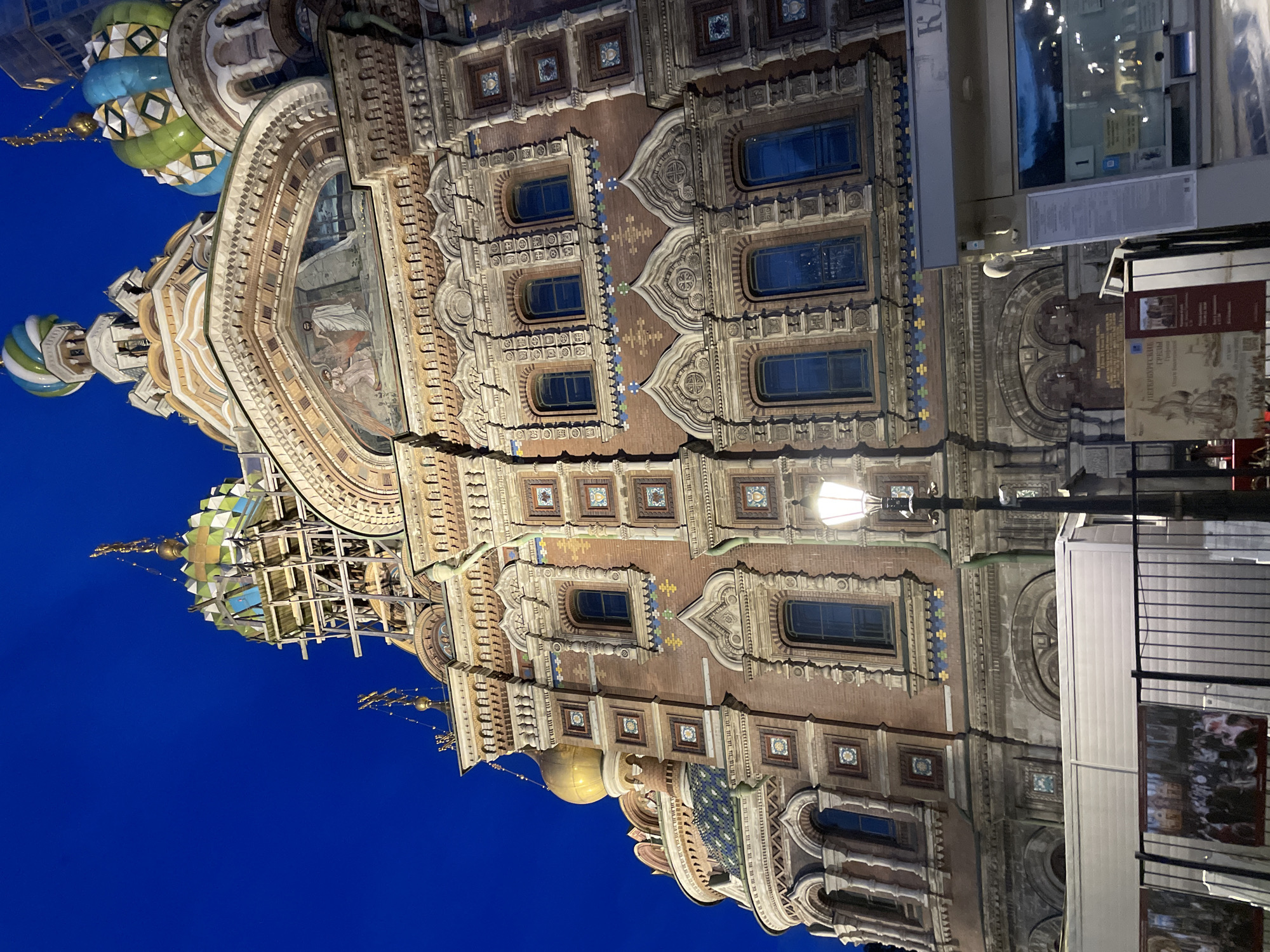 The image displays an ornately decorated building, identified as the Church of the Savior on Spilled Blood in St. Petersburg, Russia, against a deep blue night sky. The primary subject is the building's elaborate facade, viewed at an upward angle and originally oriented on its side (rotated left in the provided media).
The building features intricate architectural details, including multiple colorful onion domes. Visible domes are adorned with geometric patterns in shades of green, blue, white, and gold. The facade itself is rich with decorative elements, such as arched windows, detailed mosaic-like tile work, and sculpted stonework. One prominent archway features a fresco or large mosaic depicting figures, possibly religious scenes.
The time of day is clearly night or late dusk, indicated by the dark blue sky and the illumination of the building and a nearby street lamp. The overall atmosphere is one of grandeur and historical significance.
In the foreground on the right, parts of urban infrastructure are visible, including a black street lamp with a glowing light. Below the lamp, there are signs and display panels. One red banner clearly shows Cyrillic text including "ПЕТЕРБУРГСКИЙ" (Petersburg) and "ГЕРБАРИЙ" (Herbarium), and possibly "РУССКОЙ КУХНИ" (Russian Cuisine). Another white structure appears to be a kiosk or information stand, with visible text like "КАС" (likely part of "КАССА" for ticket office) and the number "1" on a small blue square.