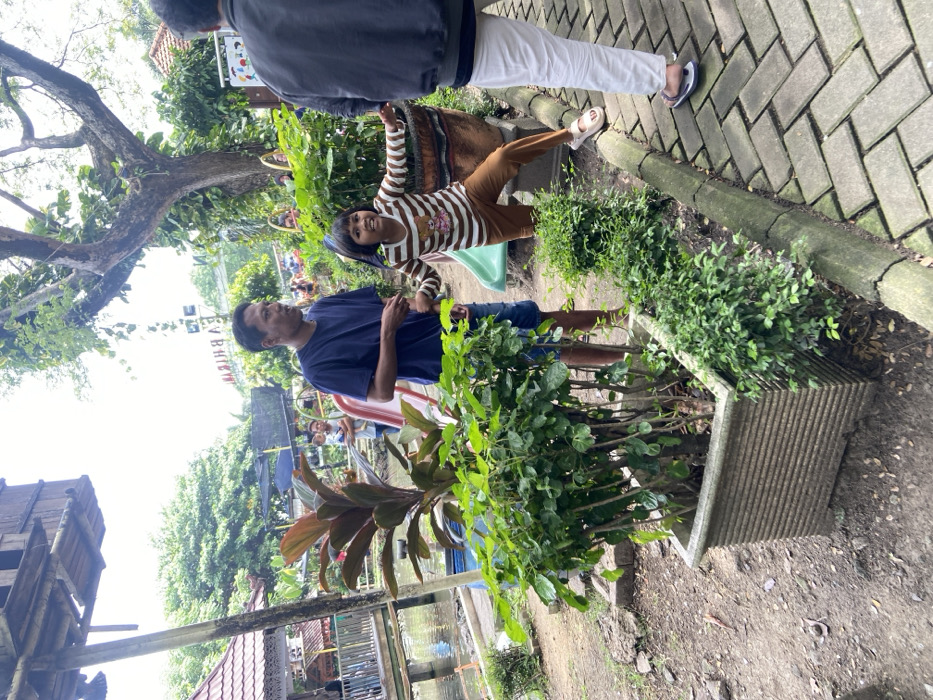 The image shows a scene in a park located in Kecamatan Sidoarjo, Indonesia at coordinates -7.441, 112.705. Three individuals are present: an adult male wearing a dark blue t-shirt and khaki shorts standing on a pathway, a young girl wearing a striped dress and brown sandals, and a second adult male wearing a long-sleeved light blue shirt and white pants. The girl is standing on a curb while gesturing to the adult male, who is looking down at her with an expression of concern. The second adult male is standing behind the girl, facing towards her. The background shows a small, wooden building with a slide and a pond. The scene is partially shaded by trees and the ground is paved with bricks. The girl appears to be engaged in a conversation with the adult male, while the second adult male watches them. A large, potted plant is located next to the curb where the girl is standing. The overall atmosphere of the image is casual and serene.