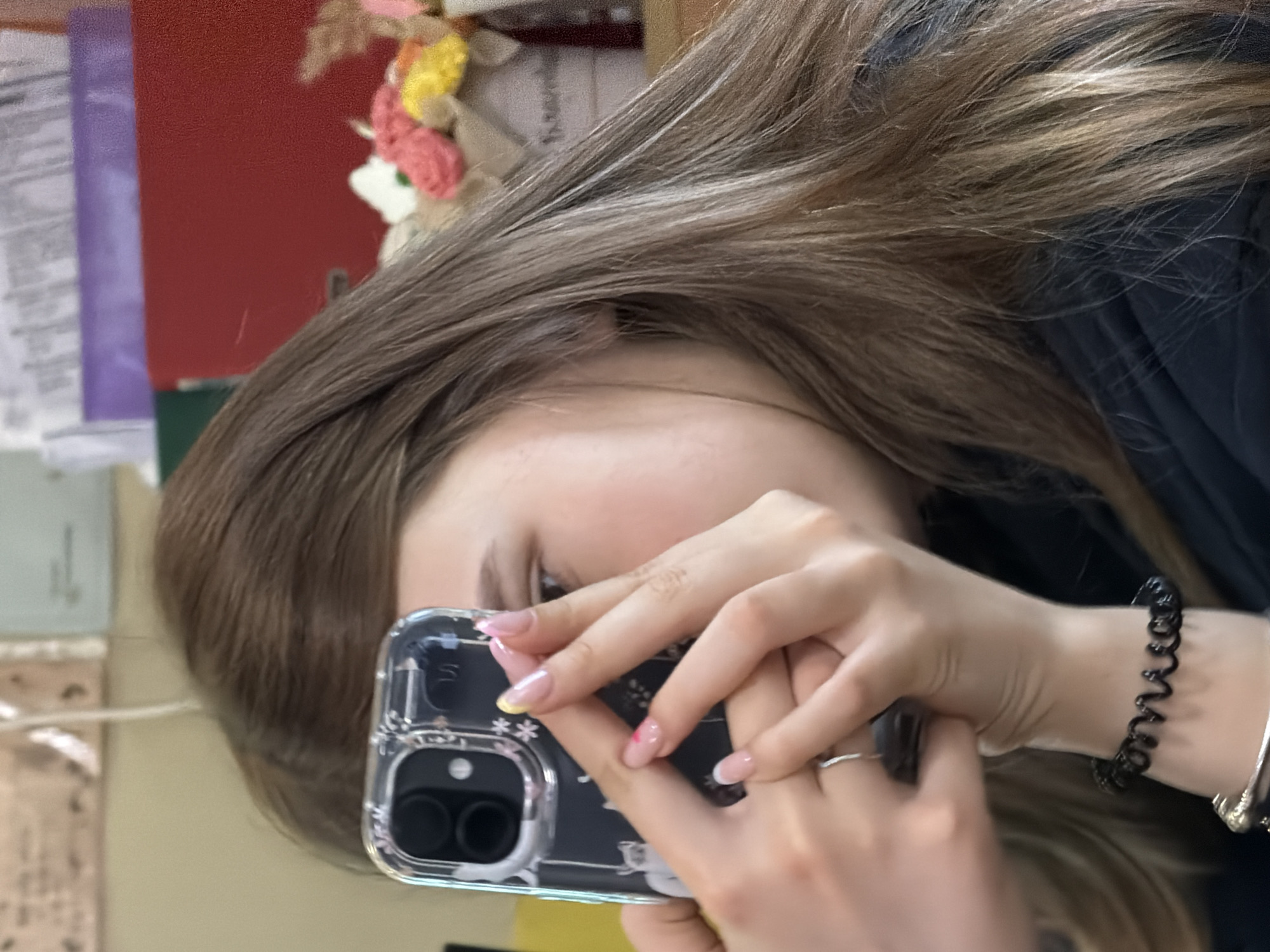 A person, identifiable by their hair, face, and hands, is holding a smartphone up to their face. The phone has a clear case decorated with floral patterns, and two prominent camera lenses. The person's nails are painted with different designs. They are wearing rings and bracelets. The background includes what appears to be a desk with documents and decorative items. The location is not discernable from the provided image. There is no verifiable evidence that this photo was taken in Panagyurishte, Bulgaria.