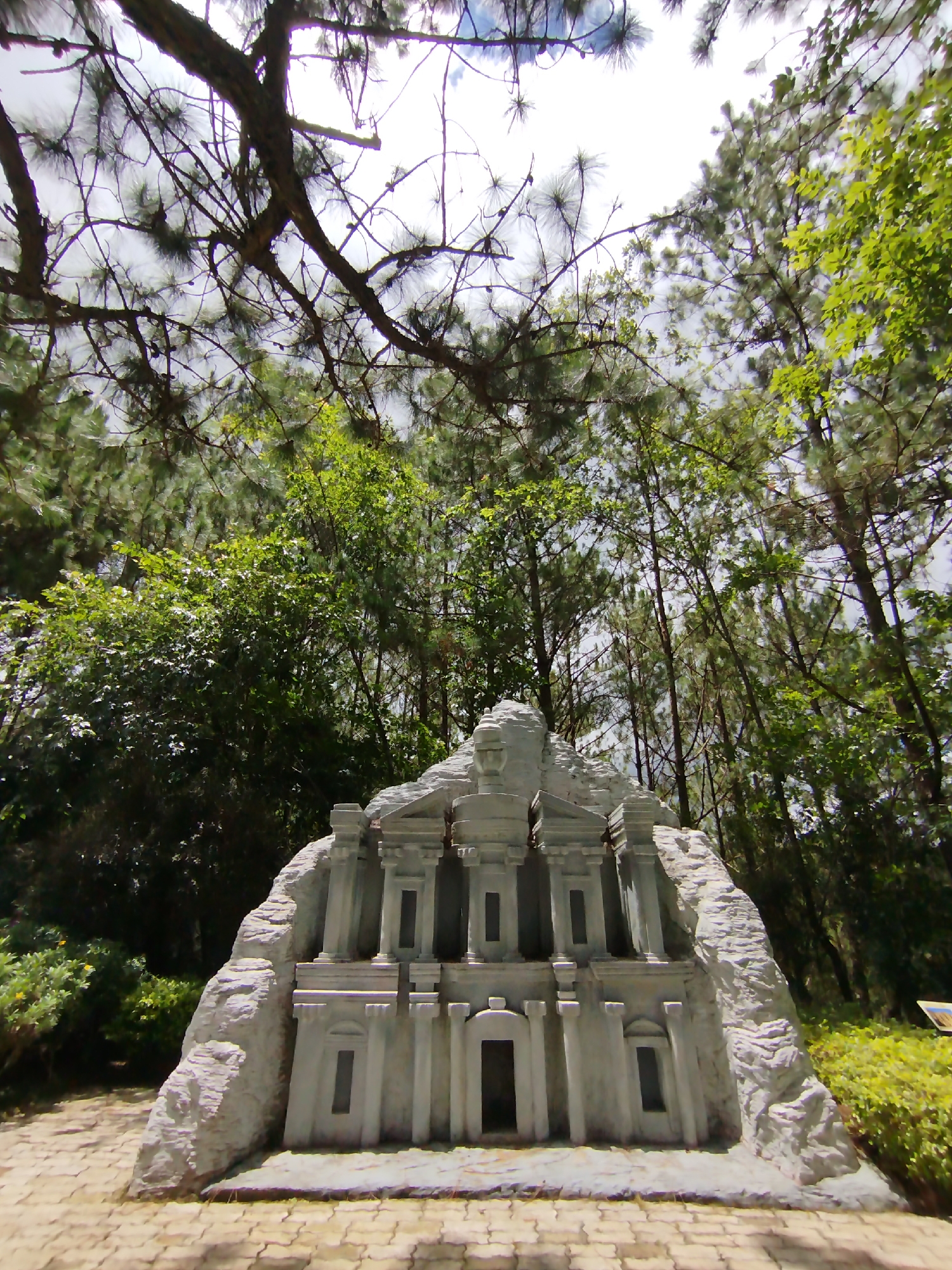 The image shows a miniature replica of the Treasury at Petra, Jordan, located in a park setting in Lam Vien - Da Lat, Vietnam. The structure is made of light grey concrete. It has a central doorway and flanking columns and decorative elements characteristic of the original monument. It is set against a backdrop of dense foliage, including various trees and bushes. A network of tree branches dominates the upper portion of the frame, partly obscuring the sky. The ground in front of the replica is paved with interlocking stone blocks.