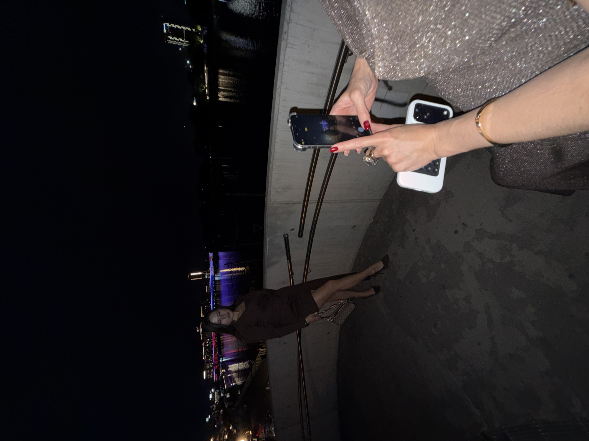 The image captures a scene at night in Belgrade, Serbia. A woman in a brown dress and heels is posing near a bridge overlooking a river and city lights. Another person is partially visible, taking a photo with two phones. The surroundings suggest an urban environment.