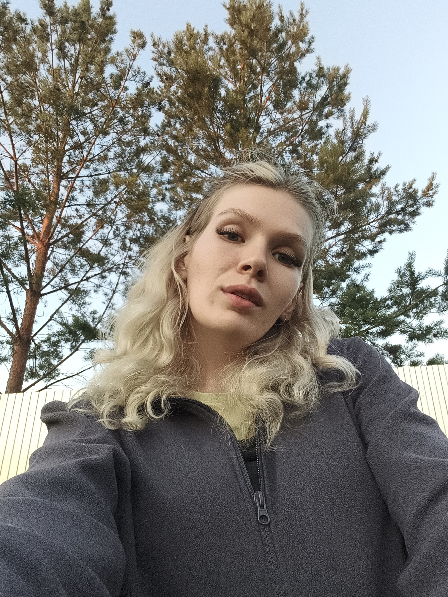 A young Caucasian woman with blonde, wavy hair is taking a selfie outdoors. She is wearing a dark grey zip-up fleece jacket over a light yellow shirt. The woman's face is tilted slightly upwards towards the camera, and she has on dramatic false eyelashes and natural-looking makeup. She appears to be looking at the camera with a neutral expression.
The background of the image features tall pine trees with scattered needles against a clear blue sky. A light-colored wooden fence is visible behind the woman, extending across the frame. The lighting suggests it is either late afternoon or early evening, with a warm glow on the pine tree on the left side of the image. The setting appears to be a natural or semi-rural outdoor area, possibly a backyard or park in Русский Вожой, Russia, given the context. There is no visible text in the image.