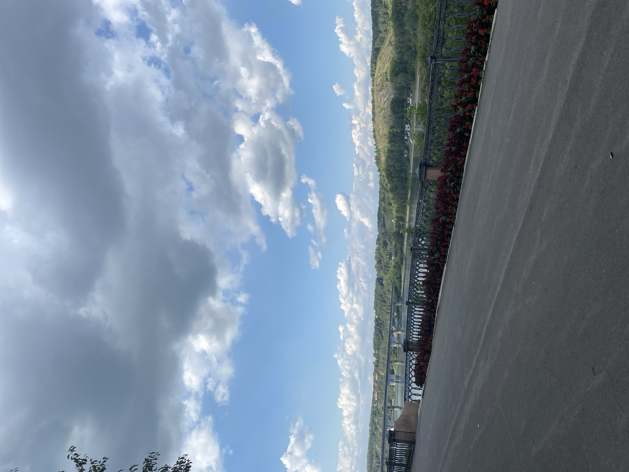 The image shows a view of the Tom River in Kemerovo, Russia, from a pedestrian walkway. The walkway is paved with asphalt and slopes downwards from right to left. Along the edge of the walkway is a decorative metal fence with red flowers planted in front of it. Beyond the fence is the Tom River, with a bridge crossing it in the distance. The far bank of the river is lined with trees, and in the background, there are hills. The sky is partly cloudy, with some dark clouds mixed with patches of blue sky. There are no people visible in the image.