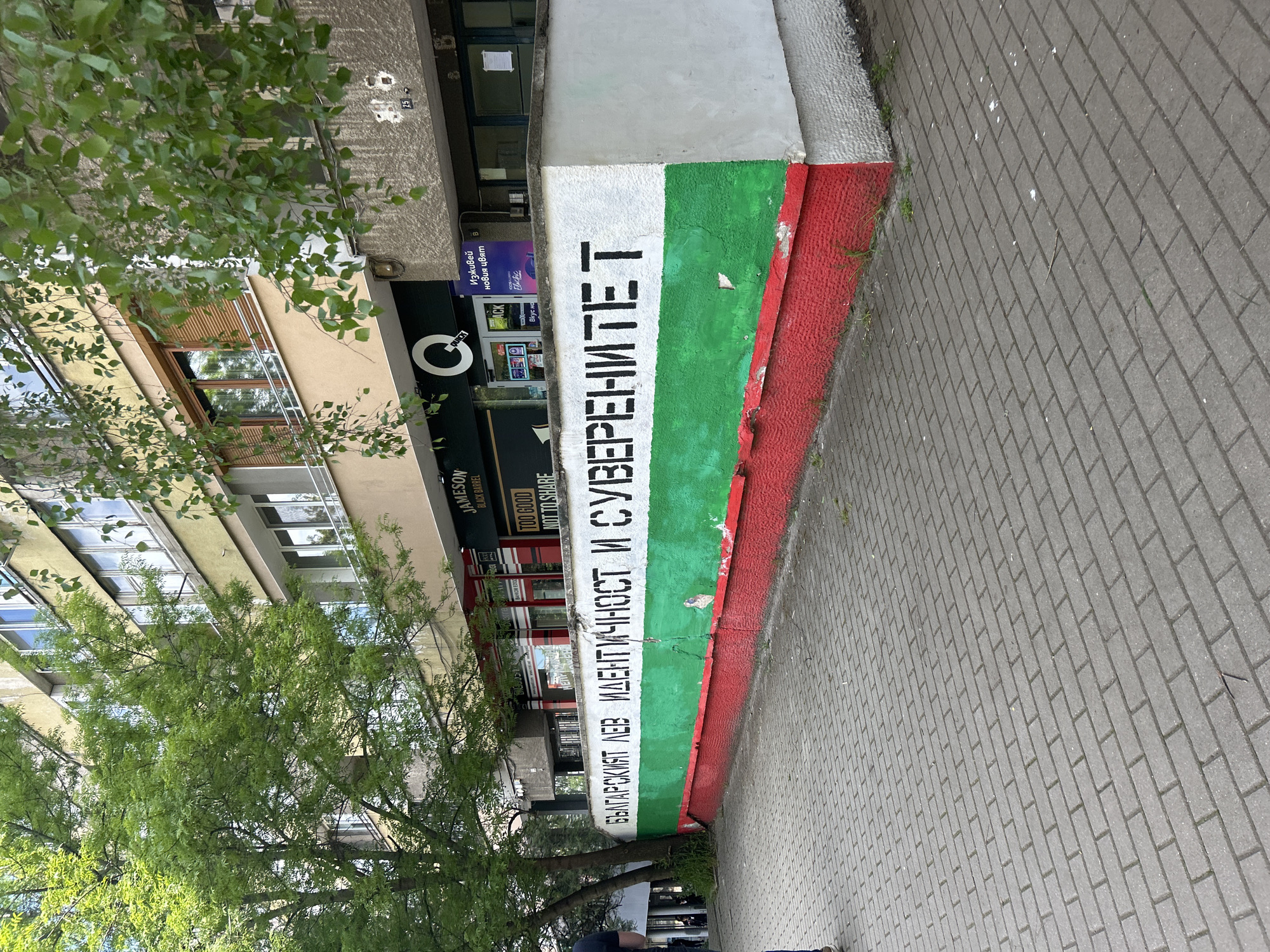 The image shows a section of a building painted with the colors of the Bulgarian flag (white, green, red). Black Cyrillic text is painted vertically over the flag. The text appears to include the words "IDENTITY" and "SOVEREIGNTY". The building also has signage for Jameson Black Barrel whisky and a store with signage that includes a stylized "Q". A tree is partially obscuring the building. The area is paved with interlocking bricks. Based on the text on the wall, the image is likely taken in Sofia, Bulgaria.
