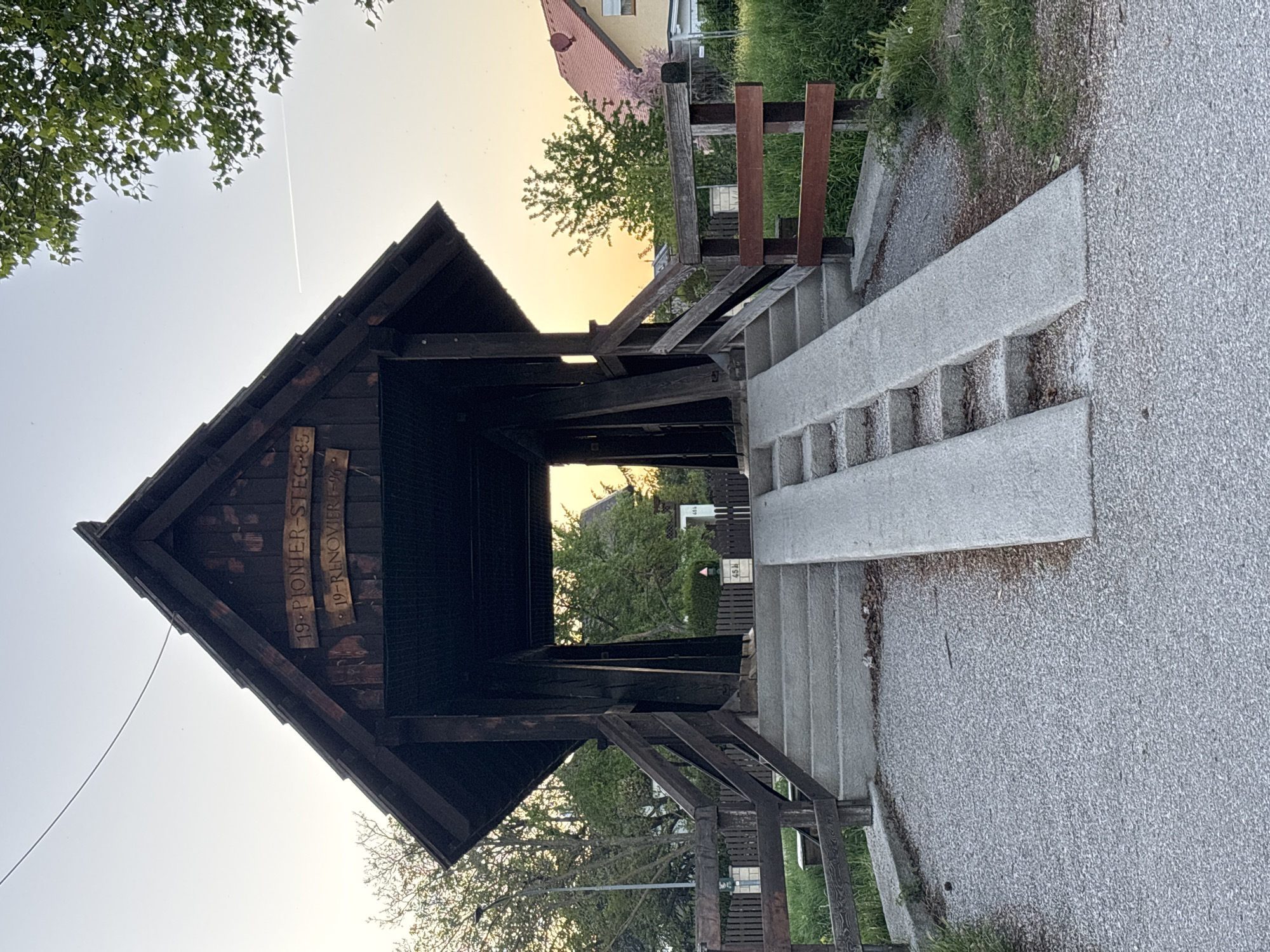 The image shows a wooden bridge structure, likely a pedestrian overpass or bridge, known as the "19. Pionier-Steg" in Wiener Neustadt, Austria. The bridge has a dark-stained wooden frame and roof, with wooden railings and supporting beams. There are concrete stairs leading up to the bridge on both sides, with concrete ramps positioned alongside the stairs. The environment includes green trees and vegetation on both sides of the bridge. A building can be seen in the background. The image is taken during sunset/dusk, with a warm glow. The path leading up to the bridge is asphalt.