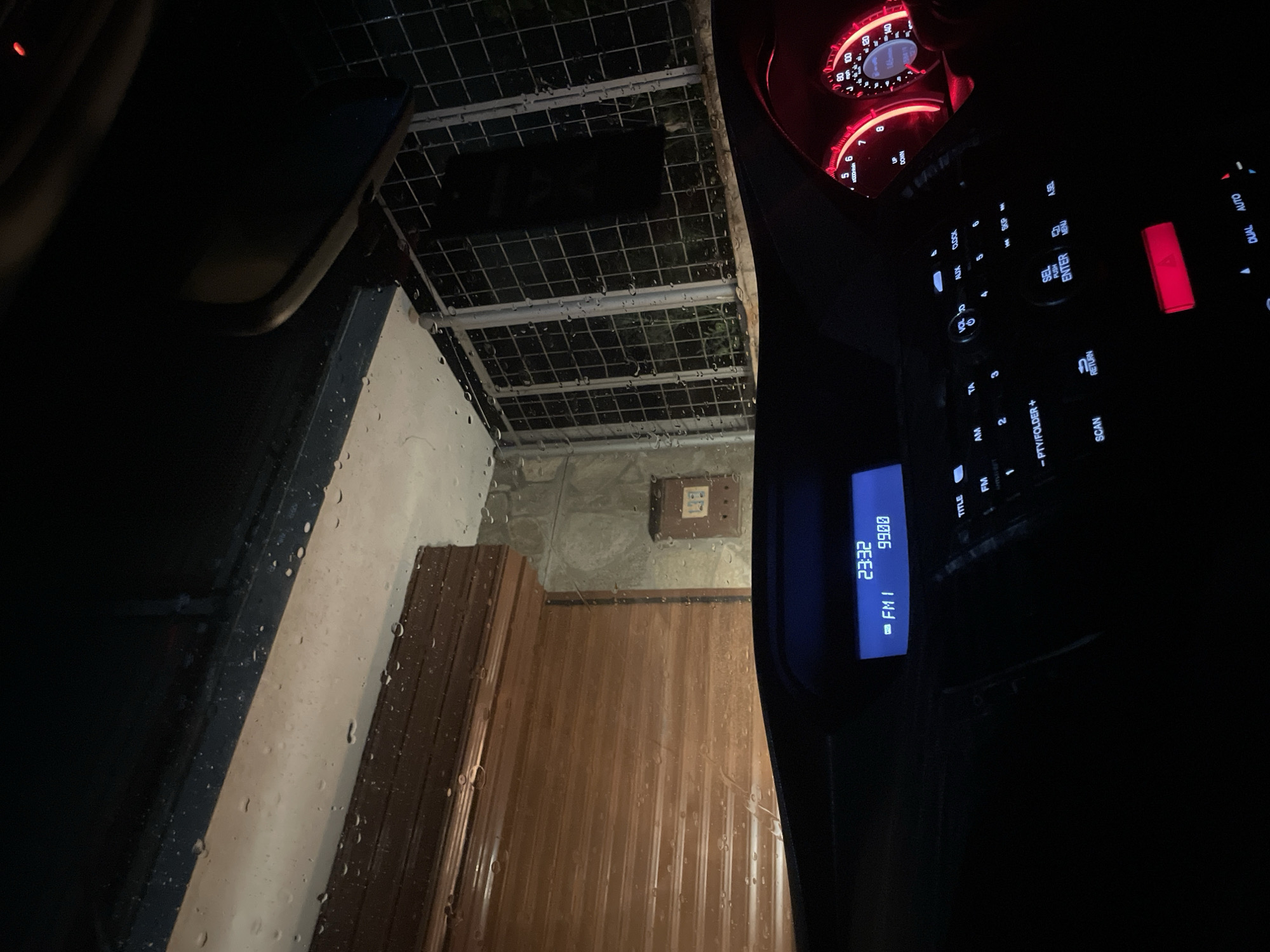 A view from inside a car, looking out at what appears to be a covered outdoor area, likely a porch or patio, on a rainy night. The car's dashboard is visible on the right, with illuminated radio display showing "23:32," the station "FM1," and the frequency "99.00." The car's speedometer and tachometer needles are barely visible, lit in red. Through the rain-streaked windshield, a white wall and a brown wooden door are discernible. The outdoor area has a metal grid roof. There is also a grey concrete wall section and a small square box on the wall. The location is indiscernible based on the provided image.
