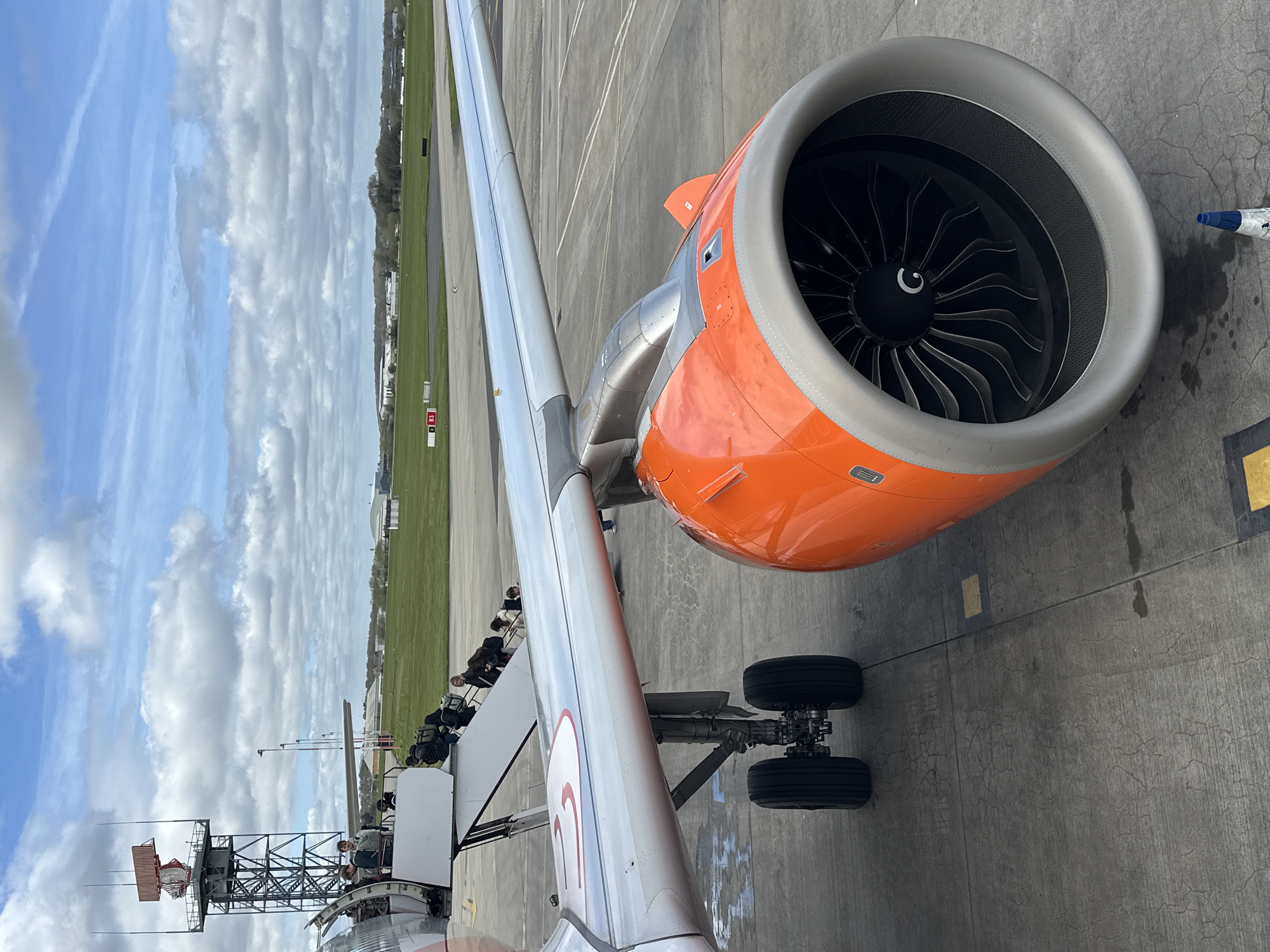 The image shows the wing and engine of an orange-and-grey aircraft on the tarmac at Southend Airport, located in Rochford, United Kingdom. Passengers are visible boarding the plane via a mobile boarding stairs connected to the wing. The sky is partly cloudy. An airport tower is also visible in the distance.