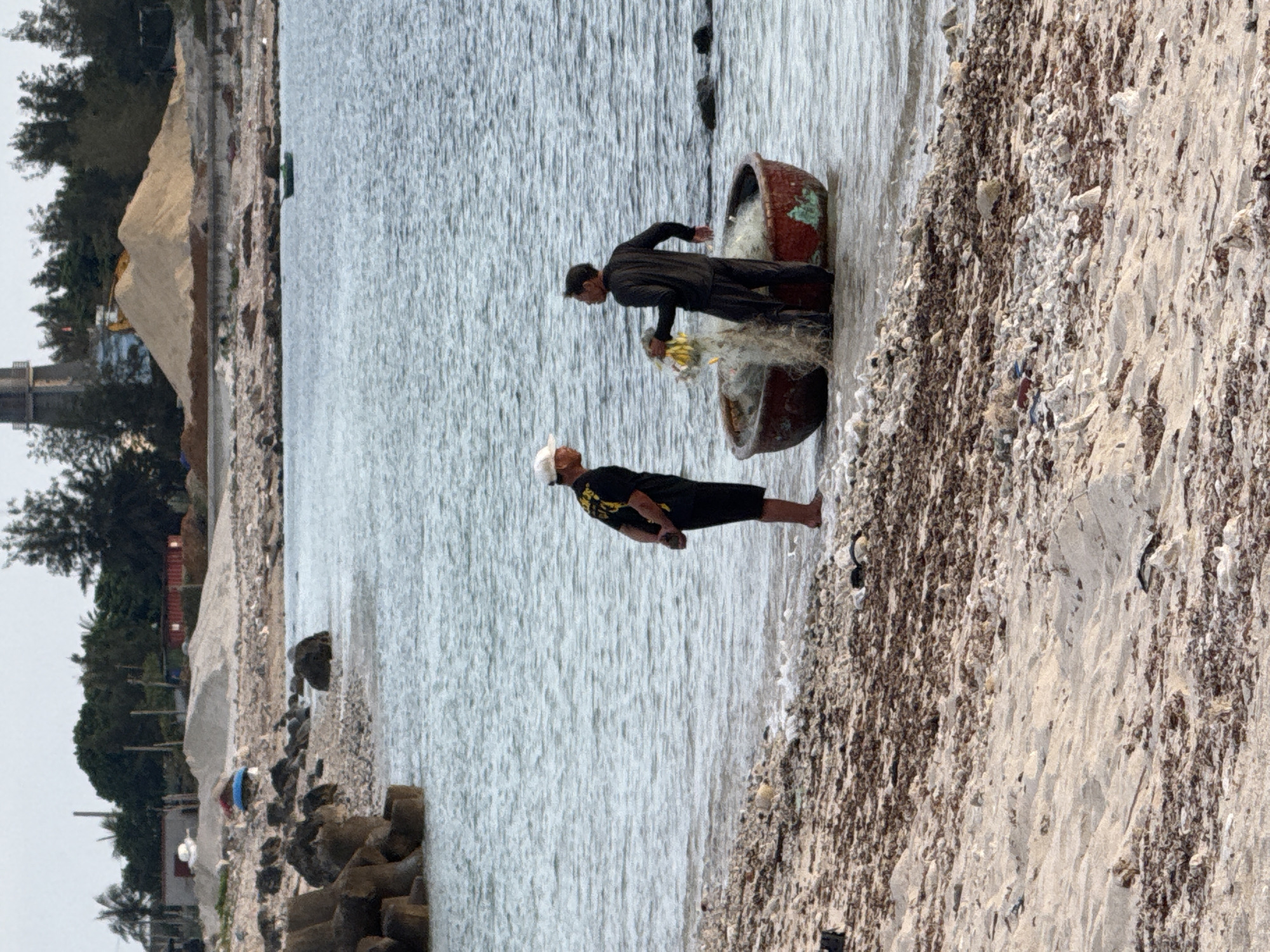 Two men stand in shallow water next to a round, red basket-like boat on the shore of Lý Sơn Island, Vietnam. The man on the left wears a black t-shirt with yellow writing and black shorts, with a white cap on his head. He faces the other man with his hands behind his back. The man on the right wears a black long-sleeved shirt and black pants. He holds a fishing net with yellow items in it, and it is draped over the boat. The boat is partially submerged in the water. The shore is a mix of sand, pebbles, and dried seaweed. In the background, a rocky breakwater made of concrete tetrapods is visible on the left, and the sea extends to the horizon. Further inland, there is a sandy hill with construction materials and buildings. The sky is overcast.