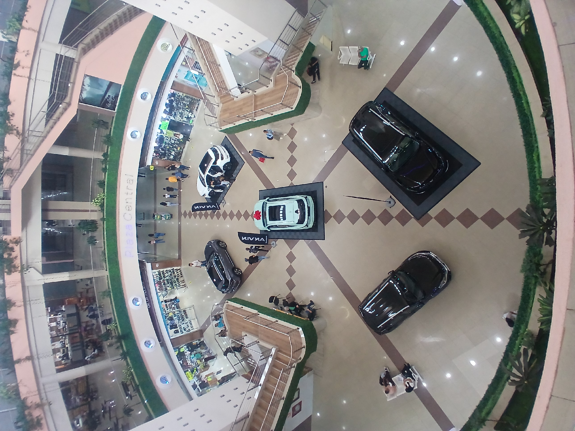 This high-angle, top-down view captures the ground floor atrium of a multi-story indoor shopping mall or commercial complex. The primary subjects are several cars on display and numerous people circulating within the space.
Four cars are prominently featured on raised platforms in the central area. Two dark-colored/black cars are on black platforms, one near the top right and another near the bottom left. A white car and a light green/teal car are displayed on adjacent lighter platforms near the center, both bearing the visible brand name "KIVNA" on a black banner.
People are engaged in various activities: some are observing the cars, others are walking along the walkways, and a few are seated on benches or chairs. The overall scene suggests a car exhibition or promotional event taking place within a busy retail environment.
Notable details include the clean, patterned floor in shades of brown and beige, the bright artificial lighting from numerous recessed ceiling lights, and the modern architectural design of the building. Curved walkways with green borders and potted plants add to the aesthetic. Shops are visible on the surrounding upper levels and along the ground floor periphery, displaying various merchandise like clothing and shoes. The word "Central" is visible on a curved wall segment on the ground floor. The atmosphere appears calm and unhurried.