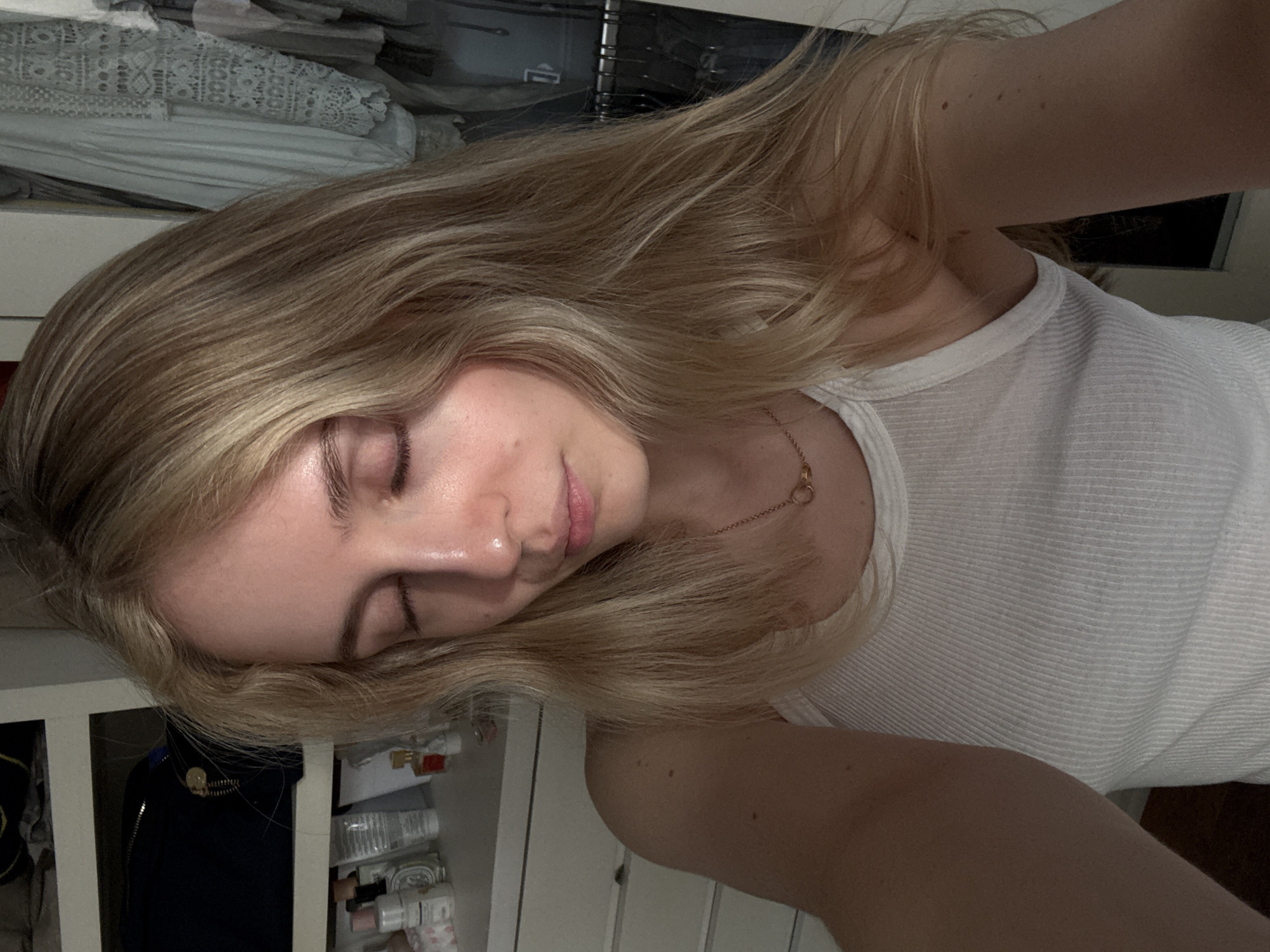 A young woman with blonde hair is lying down, possibly on a bed, with her eyes closed. She is wearing a white tank top and a delicate gold necklace. The background includes a closet or storage area with shelves containing various items, including clothing and cosmetic products. The scene appears to be indoors, possibly in a bedroom. There is no information given to suggest it is in Mexico City, Mexico.