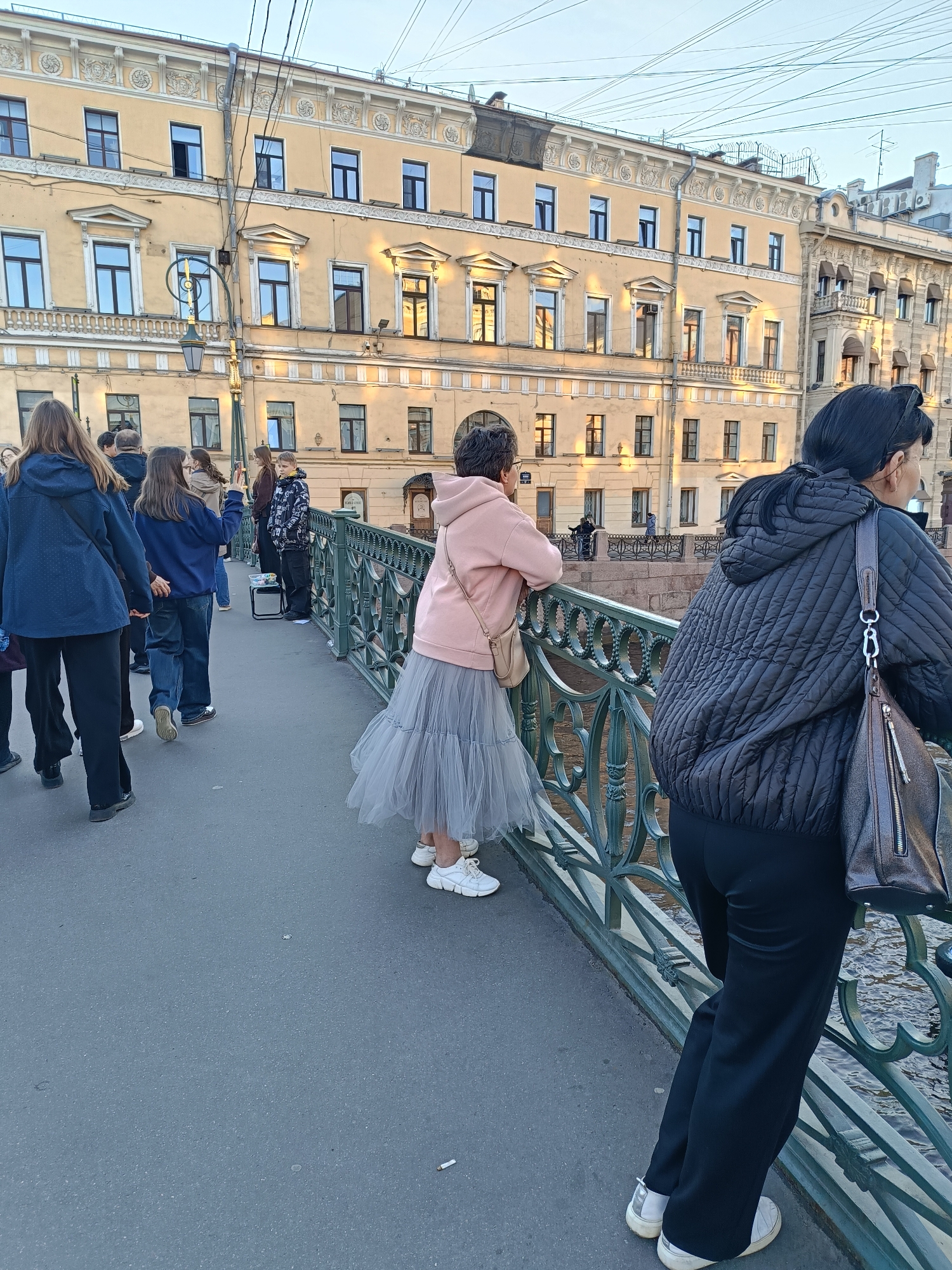 The image shows a view from a bridge in Saint Petersburg, Russia. A group of people are walking along the bridge, while two women are leaning against the bridge's green metal railing, looking out over the water. The woman on the right has dark hair pulled back and is wearing a black hooded jacket and dark pants. The woman in the middle is wearing a light pink hoodie and a gray tulle skirt with white sneakers.
Behind the people, there is a multi-story building with a yellow facade and many windows. The architectural style is characteristic of the city, with ornate detailing around the windows and roofline. A few trees are visible on the riverbank. The sky is overcast. The lighting suggests it is either early morning or late afternoon.