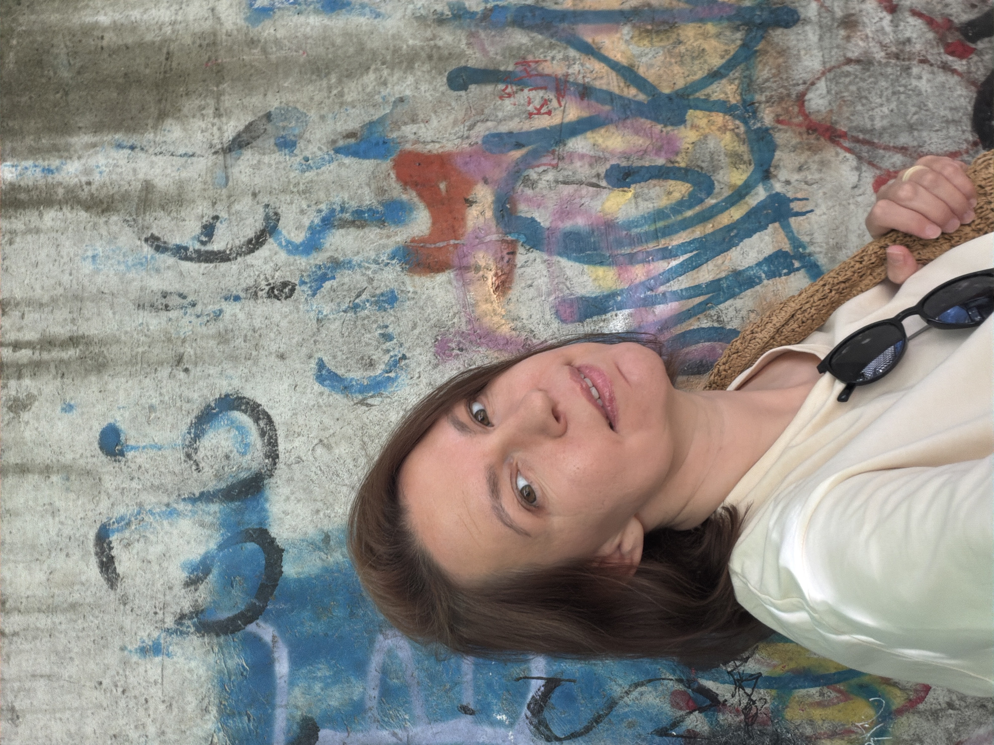 A woman is lying on a concrete surface covered in graffiti. The graffiti includes black, blue, red, yellow, and pink paint. The woman is wearing a light-colored shirt, and sunglasses are resting on her shirt. She is also holding a woven bag or hat. The background suggests an urban setting. The city is Chaikovsky, Russia.