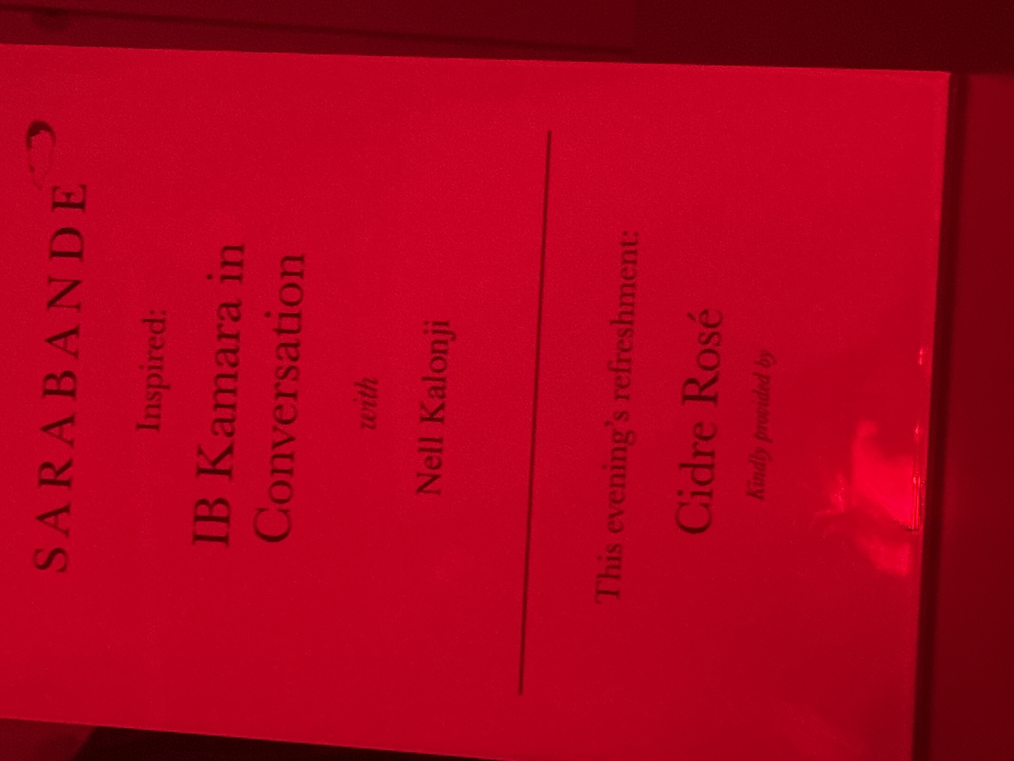 The image displays a red-lit sign or program, oriented vertically and captured at a slight angle. The dominant color is a deep red, suggesting specific event lighting, possibly in an indoor, evening setting.
The visible text on the sign, in dark contrasting font, outlines details for an event. The title or organizing entity appears to be "SARABANDE." The main event is described as "Inspired: IB Kamara in Conversation with Nell Kalonji." This suggests a discussion or talk featuring individuals named IB Kamara and Nell Kalonji, likely related to an inspiration or theme provided by Sarabande.
Below a horizontal line, further information about refreshments is provided. It states, "This evening's refreshment: Cidre Rosé." Underneath, it adds, "Kindly provided by." The "evening's refreshment" explicitly indicates the event is taking place in the evening. Given the context of London, United Kingdom, this sign is likely from a cultural event, talk, or exhibition hosted in the city. The overall presentation points to a curated and organized gathering.