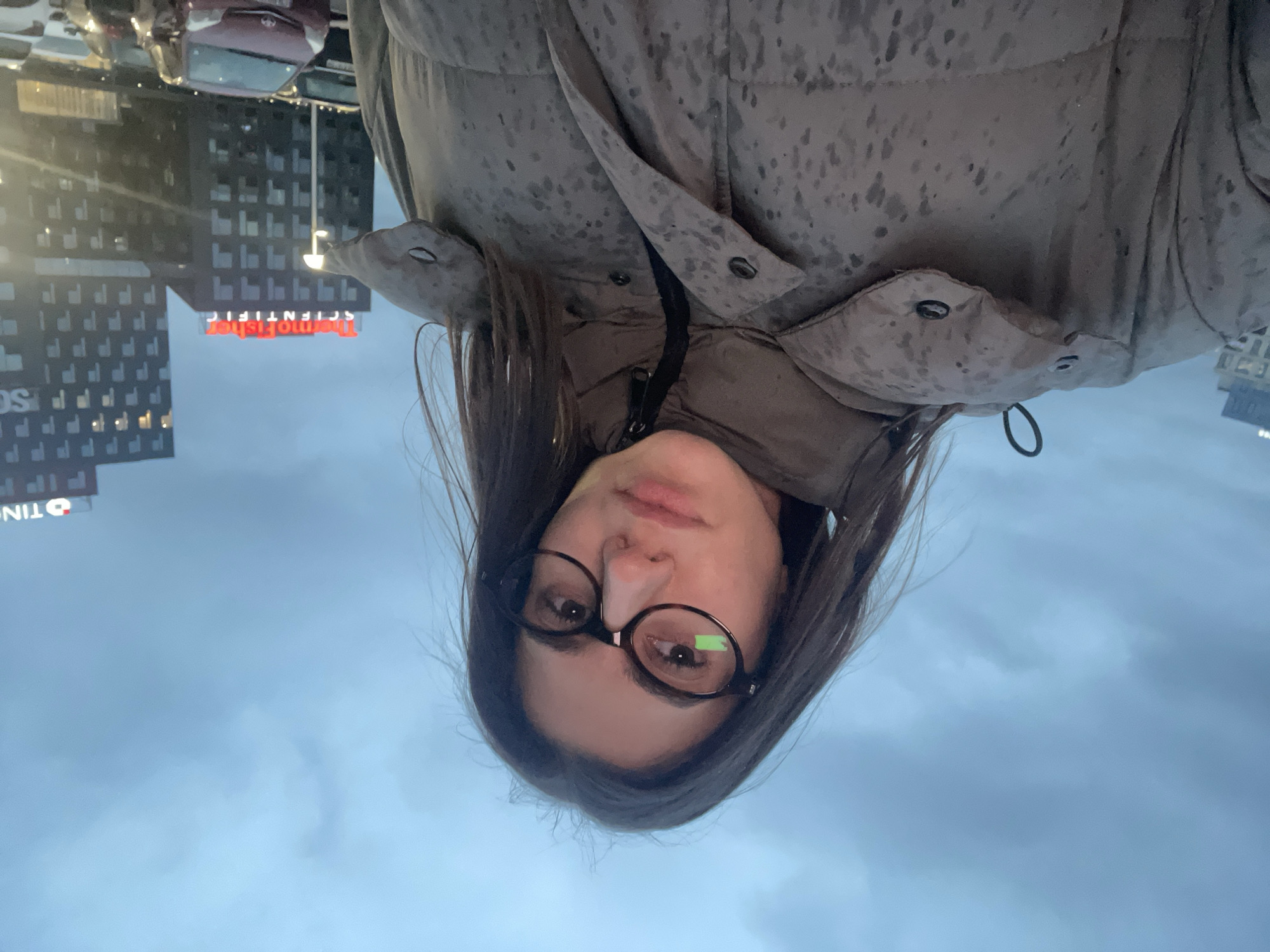 The image shows a woman wearing glasses and a puffy jacket. Buildings are visible in the background, including one with the sign "Thermo Fisher Scientific." The location is Sofia, Bulgaria. A vehicle is parked nearby. The sky is overcast.