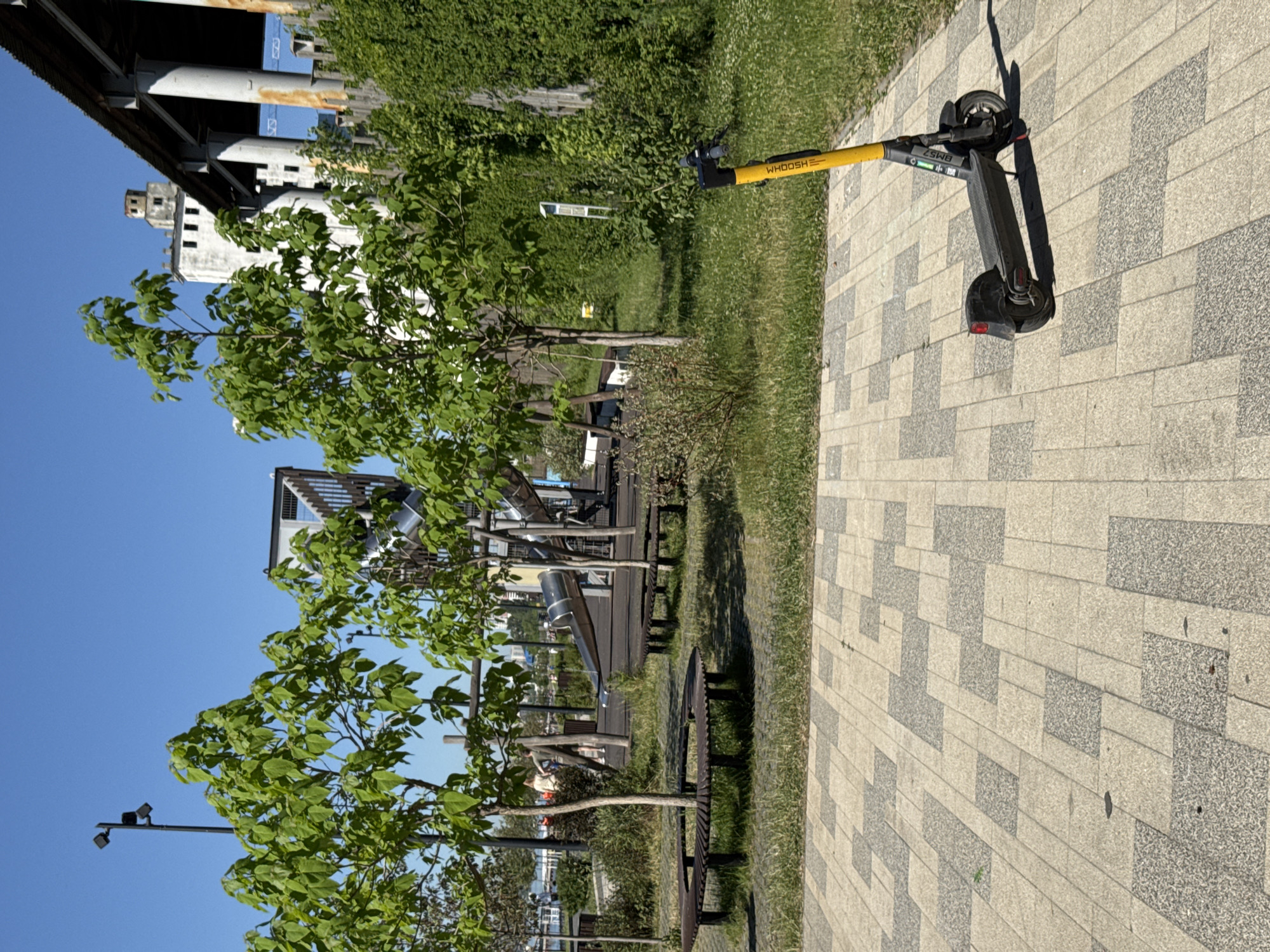 Here's a description of the image:
The image shows an outdoor scene, likely a park or recreational area in Saratov, Russia, on a sunny day. The sky is clear and blue.
In the foreground, a "WHOOSH" branded electric scooter is parked on a paved pathway. The pathway consists of alternating light and dark gray rectangular pavers. The scooter is black and yellow. To the left of the pathway, there is a grassy area with trees and benches. Some of the trees are young, with vibrant green leaves.
Further in the background, there is a structure with a modern design, possibly a pavilion or an architectural feature. A bridge or overpass is visible in the upper left corner.