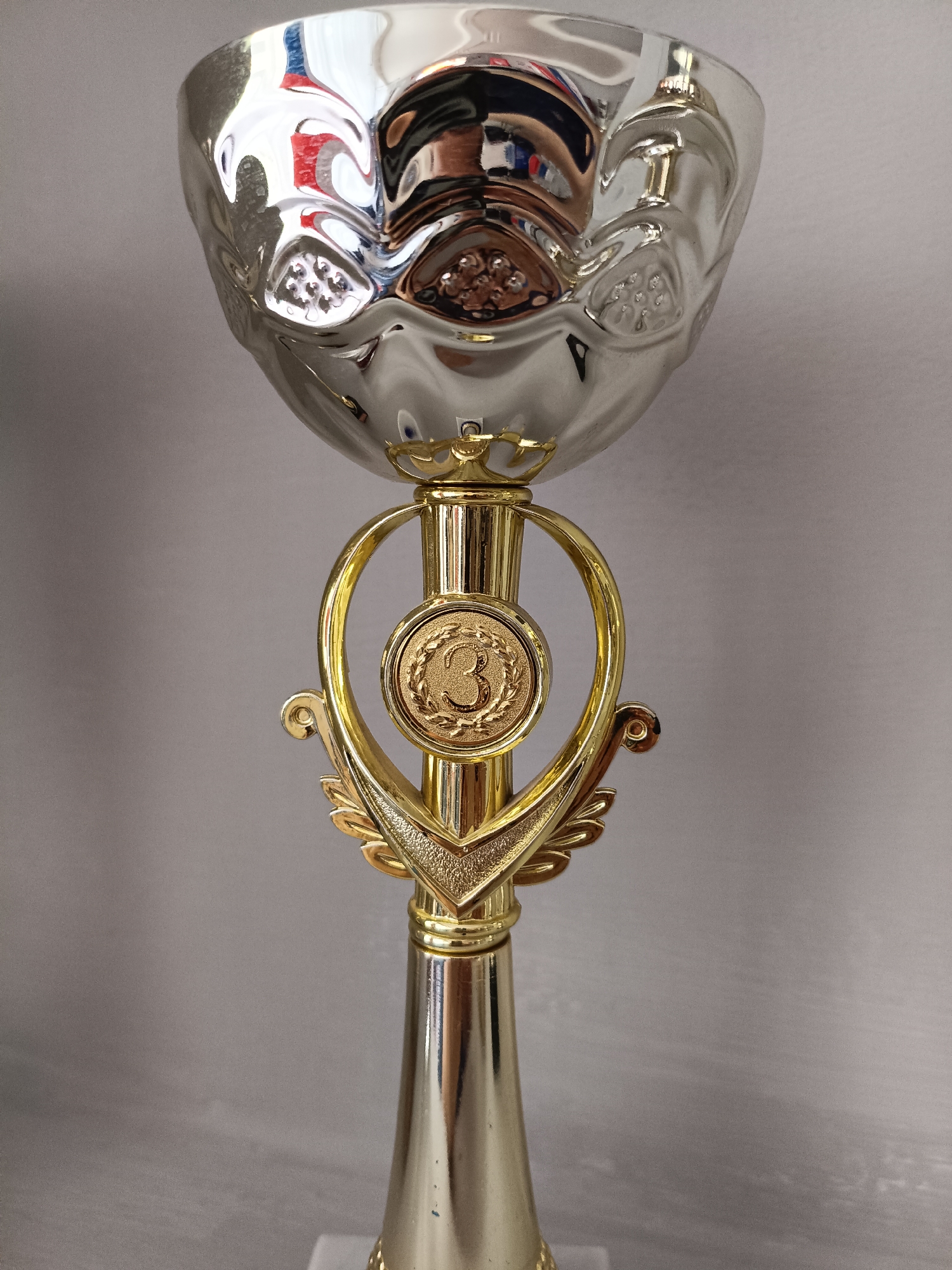 This is a close-up, vertical, eye-level shot of a silver trophy with gold accents. The trophy is predominantly silver and has a bulbous, textured cup at the top. Below the cup is a gold stem with ornate detailing. A gold, heart-shaped element encircles a circular medal. This medal features a gold embossed number "3" surrounded by laurel leaves. The trophy is set against a plain, light gray background with a subtle texture, suggesting an indoor setting. The lighting appears to be neutral, without strong shadows or specific time-of-day cues. There is no discernible activity or event taking place, as the trophy is stationary. The focus is entirely on the trophy itself, highlighting its metallic sheen and decorative elements. The overall impression is one of award or achievement.