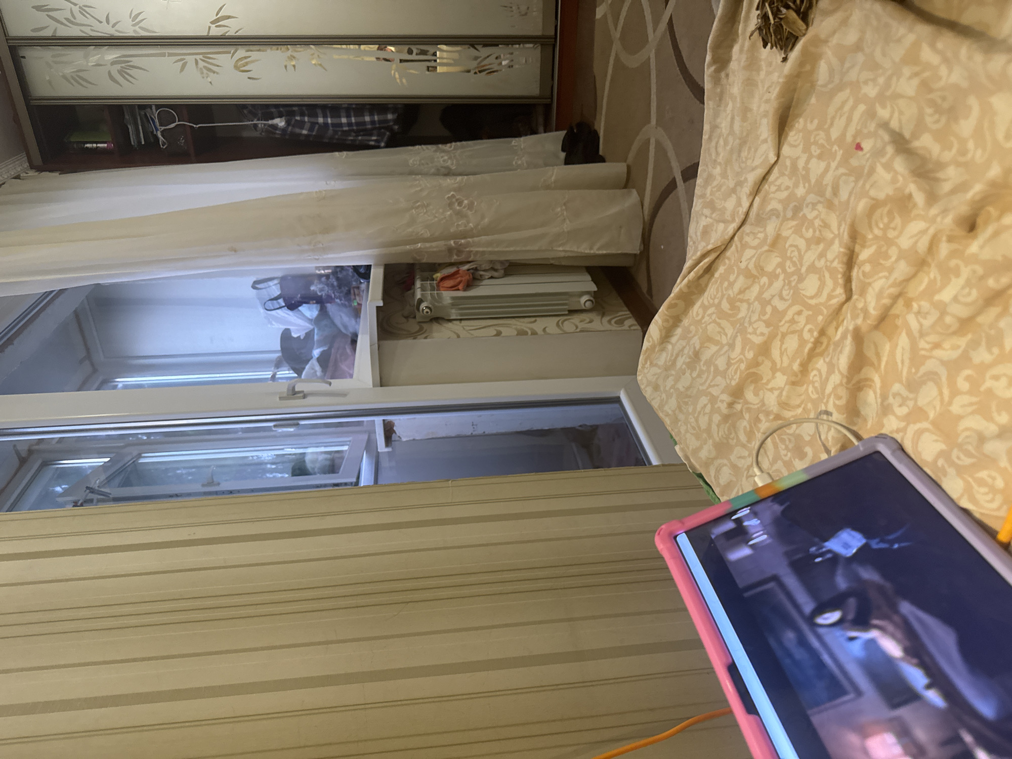 The image shows an indoor scene, likely a bedroom. In the foreground, a tablet computer with a pink and purple case is resting on a patterned bedspread. The tablet screen displays an image of what appears to be a person. To the left of the tablet is an orange cable. Behind the bed, there is a window with a view of what looks like a balcony. On the balcony, there are several bags and items. To the right of the window is a white curtain. Further to the right, a large wardrobe with mirrored doors, decorated with a bamboo pattern, is visible. Inside the wardrobe, clothes are hanging. In the corner, near the window, is a white radiator with some clothes draped over it. The floor is covered with a patterned carpet. The scene is illuminated by artificial light.