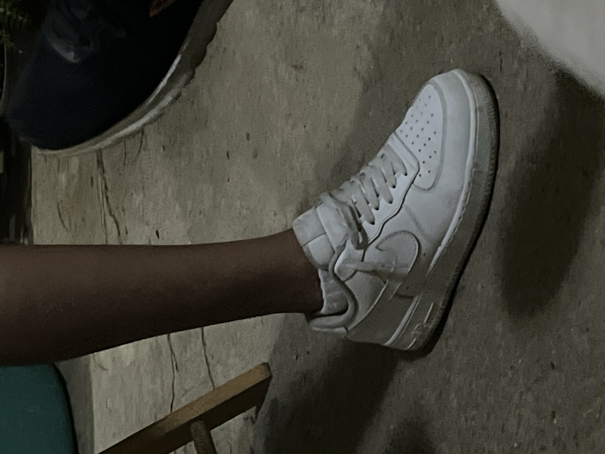 The image shows a close-up of a person's right leg and foot, wearing a white Nike Air Force 1 sneaker. The person is standing on a cracked concrete surface, possibly a sidewalk or outdoor patio. A portion of a wooden chair is visible to the left of the leg. In the background, on the left, a turquoise cylindrical object and on the right, a dark shoe are partially visible. The lighting suggests it is nighttime or a dimly lit indoor space. The image's composition focuses on the sneaker and the lower leg, with the surroundings providing context. The specific location in Santa Ana, Venezuela, is not determinable from the visual information provided in the image.