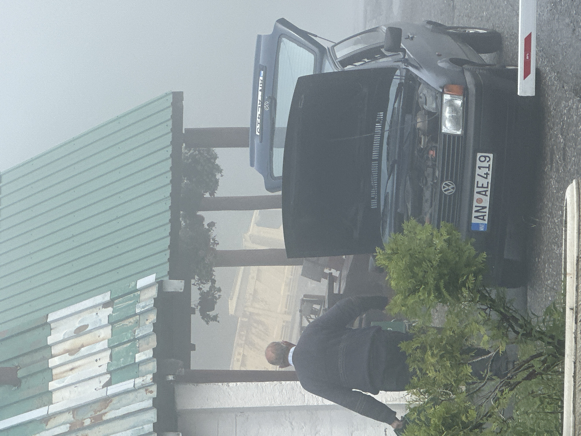 In Trešnjevik, Montenegro, amidst a dense fog, an older man wearing a navy blue sweater and dark pants stands near a dark-colored Volkswagen hatchback. The car's hood is open, revealing its engine. The rear hatch of the car is also open. A license plate on the front of the car reads "AN AE 419". The scene takes place outdoors, with a building featuring a green corrugated metal roof to the left. Vegetation, including bushes and trees, is visible in the foreground. The overall atmosphere is hazy and obscured by fog.