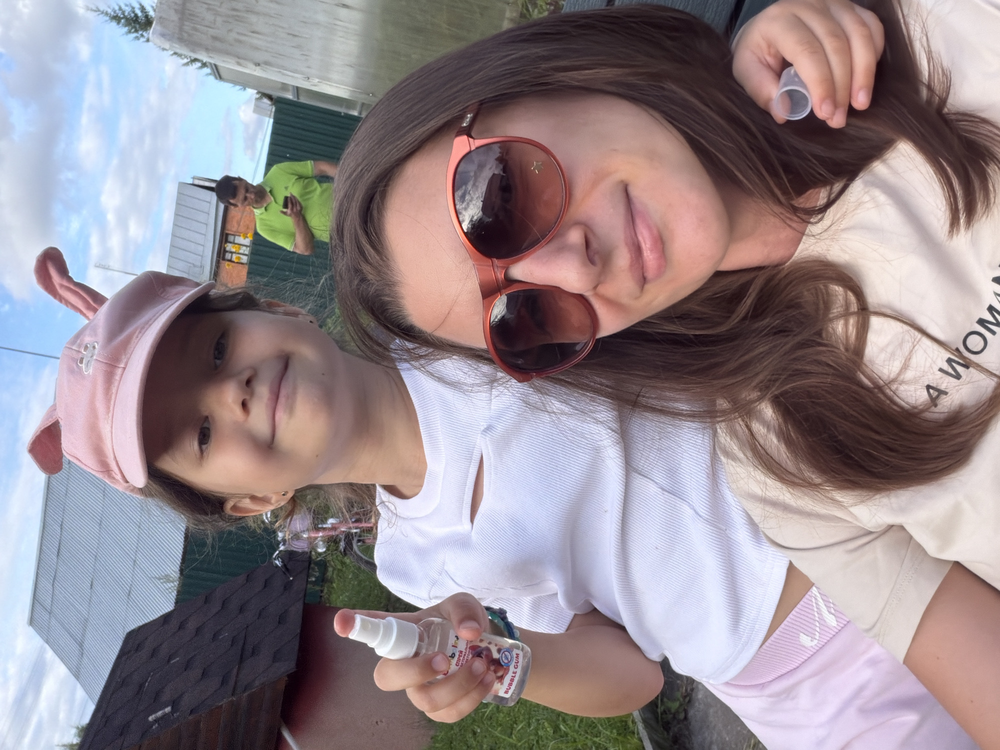 In the city of Nizinka SNT, Russia, a young girl with a pink hat adorned with bunny ears is taking a selfie outdoors. She is holding a small bottle of bubble gum scented spray and is positioned in front of an adult woman wearing sunglasses and a light-colored t-shirt. In the background, a man in a green shirt is visible, looking at his phone. The scene appears to be in a backyard or a rural setting, with houses and trees visible.