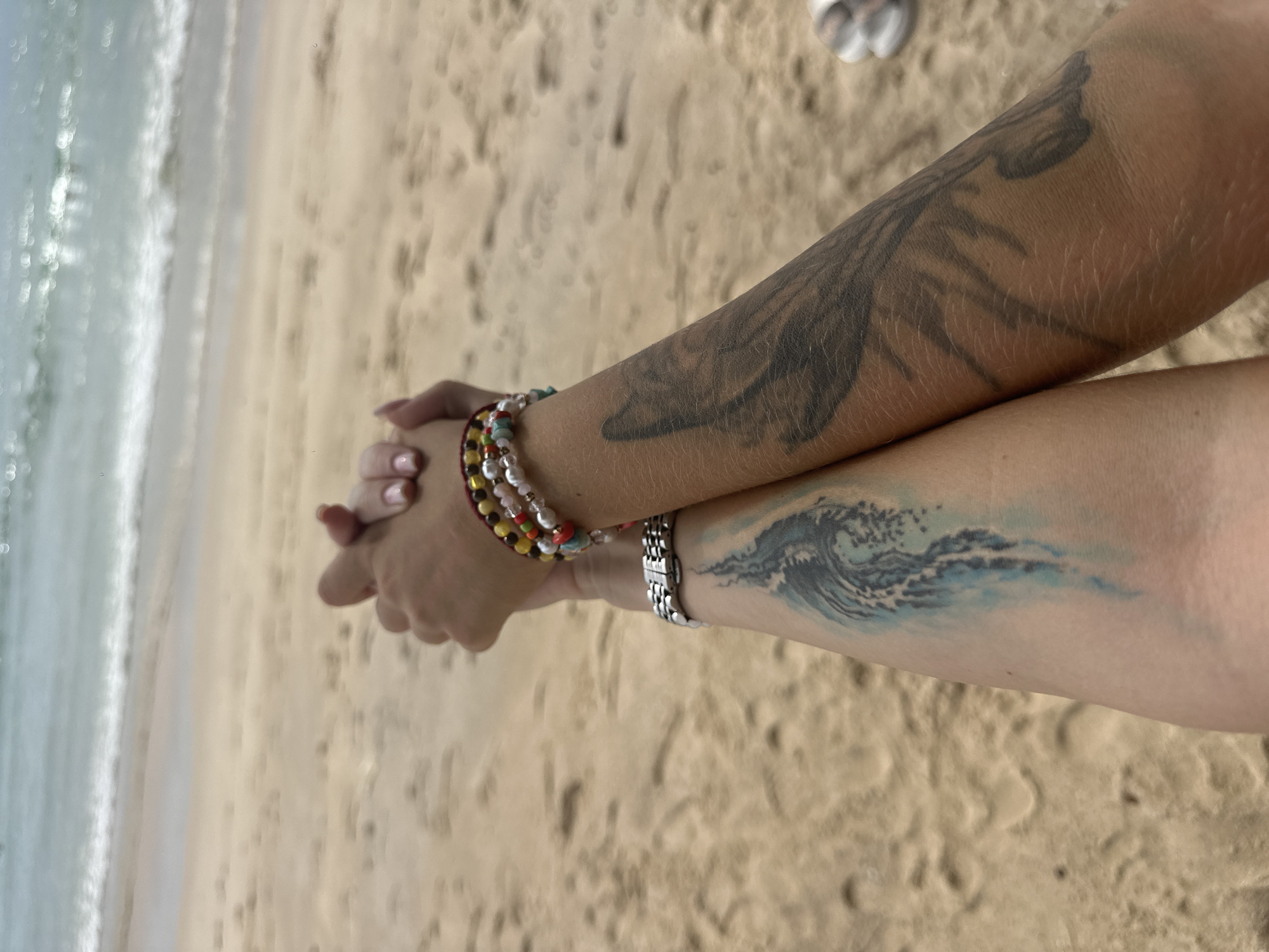 This media file shows a close-up of two arms on a sandy beach, with the ocean in the background. The arms are interlocked, suggesting a close relationship between the individuals. One arm has a dark, stylized tattoo that appears to be a wing or feather, while the other arm features a blue watercolor-style tattoo of a breaking wave. Both arms are adorned with bracelets. The left arm has a beaded bracelet with colorful beads and pearls. The right arm sports a metallic, link-style watch.
The background consists of a sandy beach and the ocean's edge, with gentle waves lapping the shore. The sand is light brown, and the water is a pale blue-green. The lighting suggests it is daytime, possibly a sunny day, given the bright sand and the water's appearance. The overall mood conveyed is one of relaxation and connection, likely at a beach vacation or outing. There is no visible text in the image.