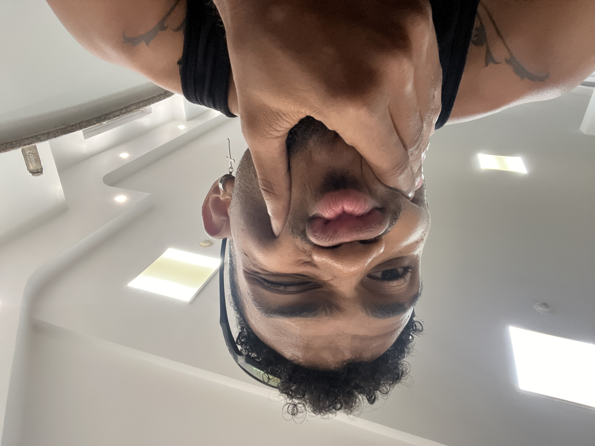 The image is an upside-down selfie of a person with medium brown skin. The person is wearing a black tank top and sunglasses on top of their head. They have short curly black hair, and the skin on their face appears sweaty. The person's right hand is around their face, distorting their mouth. An earring dangles from their left ear. The background is a white ceiling with recessed lighting and a light fixture. The location is undetermined.