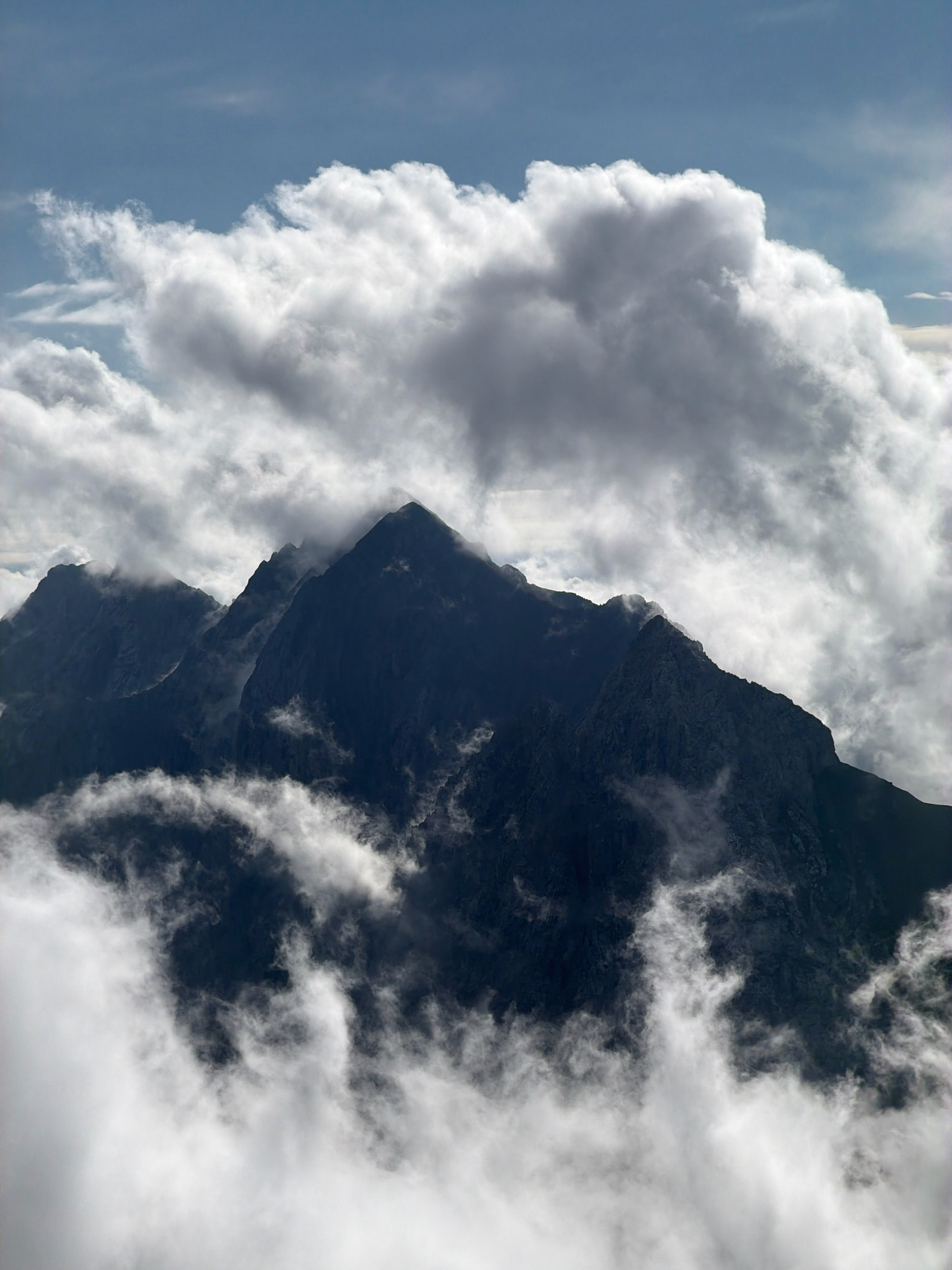 This is an image of a mountain range partially obscured by clouds, taken from a high vantage point. The peaks are dark and jagged, silhouetted against a bright, cloudy sky. The clouds are thick and white, some appearing fluffy and others more wispy and diffused. The atmosphere appears to be dynamic, with cloud cover varying across the scene, creating a sense of depth and drama. There are no people, animals, or man-made structures visible in this image. The natural environment of the mountains and clouds dominates the composition. The location is likely the Bavarian Alps, as the question specifies Garmisch-Partenkirchen, Germany, which is situated in this region and known for its mountainous terrain.