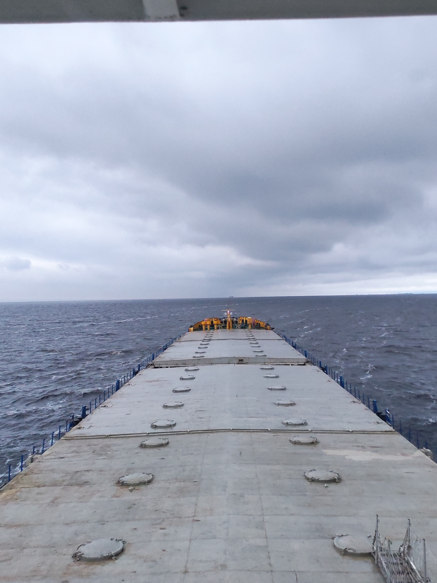 View from the deck of a ship at sea under a cloudy sky. The deck is gray and has a series of circular hatches. The sea is dark and choppy. The location is Unknown, Unknown.