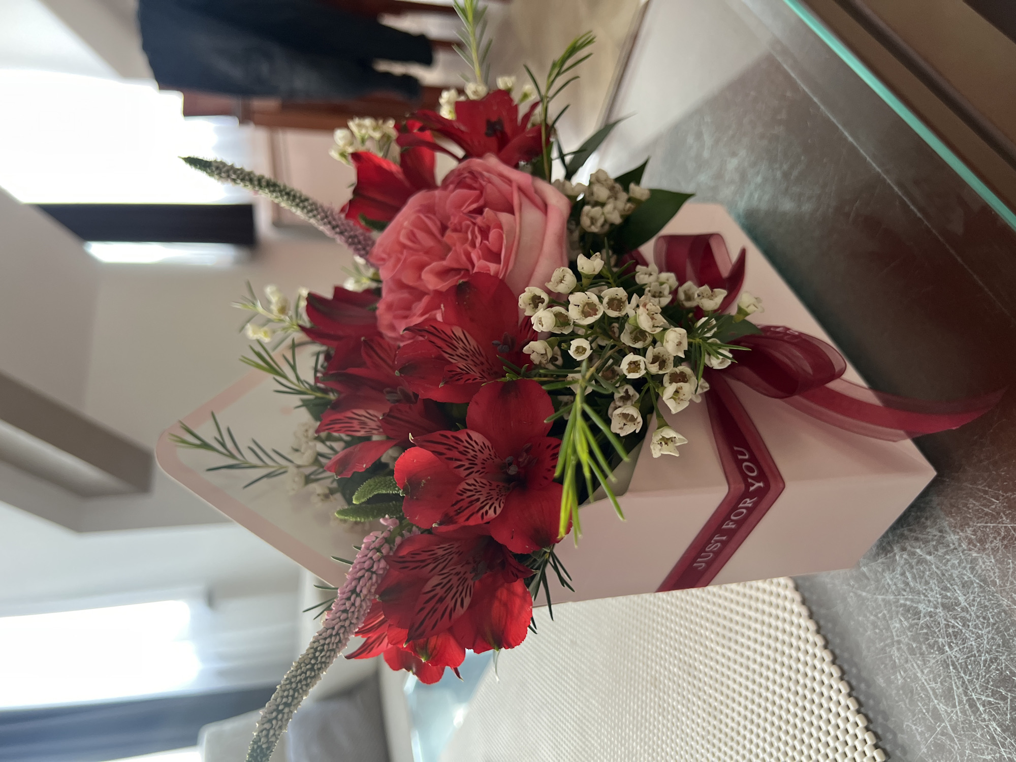 A bouquet of flowers in a light pink, hexagon-shaped box with the words "JUST FOR YOU" printed on a dark red ribbon that wraps around it sits on a grey patterned surface. The bouquet includes red Alstroemeria flowers with dark specks, a pink rose, small white flowers, and greenery. The surface appears to be a table or countertop. In the background, blurred elements of a room can be seen, including furniture and parts of a window. The location in Varna, Bulgaria is not known from the image.
