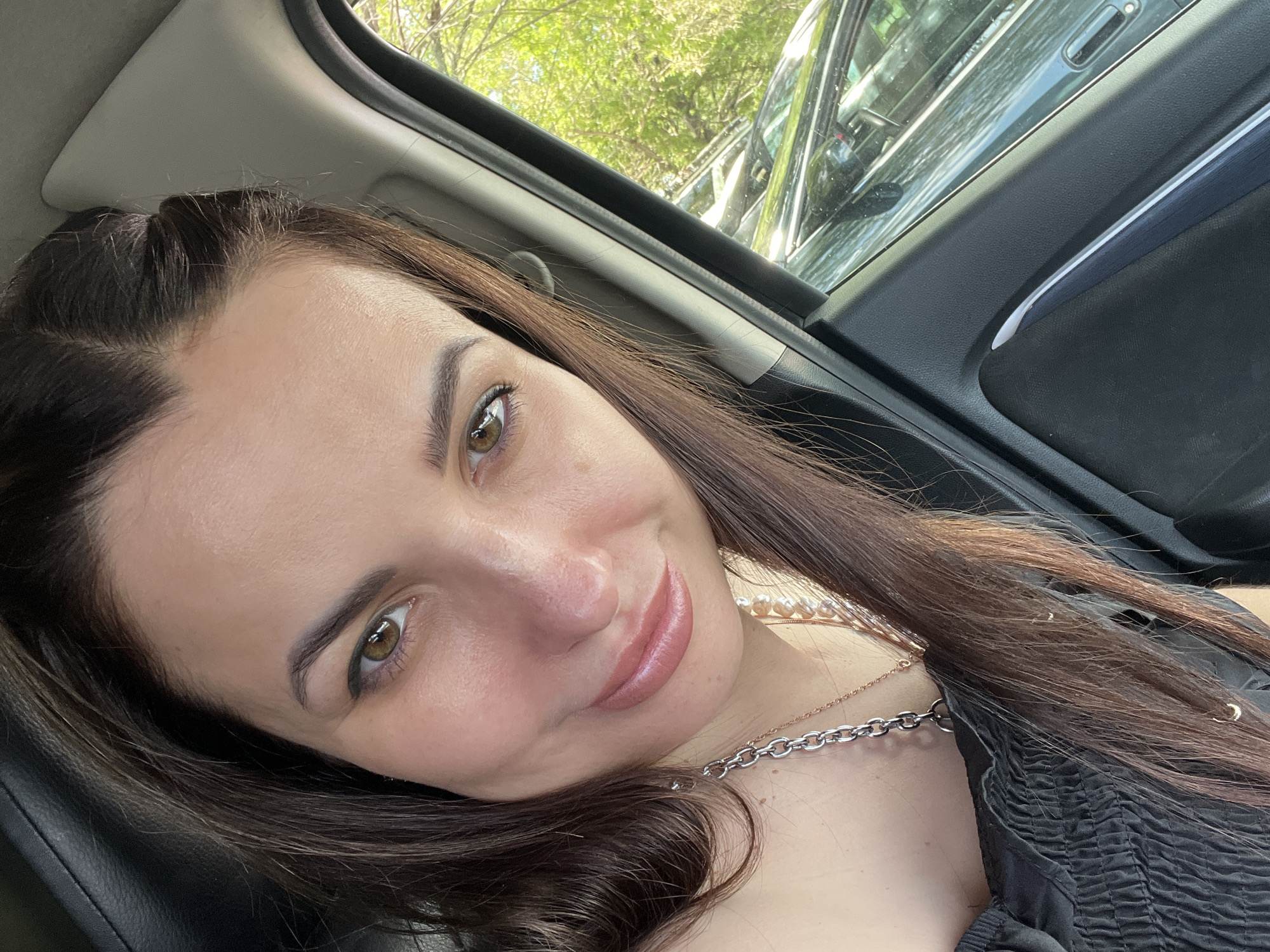 The media file features a close-up selfie of a young woman, taken from a slightly high angle, with her head tilted, looking directly at the camera. She has long, dark brown hair that cascades over her shoulders and chest, showing hints of lighter brown or reddish tones in the longer strands. Her eyebrows are dark and neatly shaped, complementing her striking light brown or amber eyes, which are subtly lined with eyeliner. Her lips are full, adorned with a light pink or nude gloss, and she wears a gentle, serene smile, conveying a sense of calm and pleasantness.
She is dressed in a black top with intricate ruched detailing on the chest. Adorning her neck are two necklaces: a prominent silver-colored chain with chunky links, and a finer, gold-colored chain layered beneath what appears to be a string of pearls.
The setting places her inside a vehicle, likely a car, given the visible interior elements such as the door frame, window molding, and parts of the headliner. Through the car window, vibrant green foliage from trees is visible, along with reflections that suggest other vehicles or structures, indicating an outdoor environment during the day. The scene is brightly lit by natural sunlight, suggesting a clear and sunny day. There is no visible text in the image.