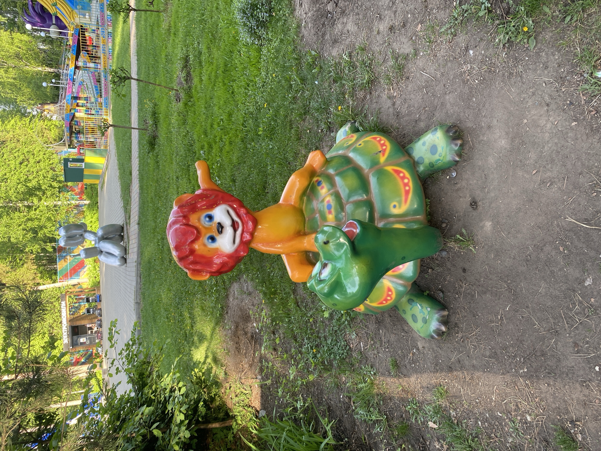 The image shows a children's playground in Izhevsk, Russia. The main subject is a brightly colored sculpture depicting a lion riding a turtle, with a crocodile head emerging from the turtle's shell. The sculpture is placed on a patch of grass adjacent to a dirt path. In the background, there is a paved walkway, green grass, and trees. Further in the background, amusement park rides and structures are visible. To the left, there are gray animal sculptures.
