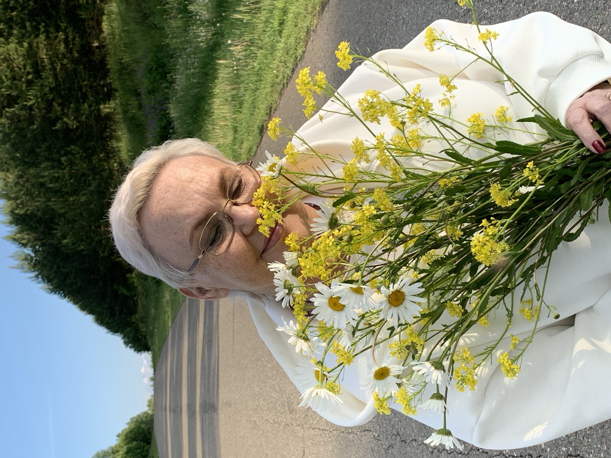 An older woman, with short, light-colored hair and round eyeglasses, is depicted outdoors in bright daylight, likely in Bol'shoi Bukor, Russia. She wears a light-colored, possibly white or off-white, hooded top. Her left hand, adorned with dark red nail polish, holds a large, vibrant bouquet of wildflowers. The bouquet predominantly features numerous small yellow blossoms and several white daisies with yellow centers, some of which partially obscure her face.
The woman gazes gently towards the camera, conveying a serene expression. The background reveals a paved path or road, flanked by lush green grass and dense trees under a clear blue sky. This setting suggests a natural or park-like environment. No visible text is present. The scene evokes a sense of tranquility and an appreciation for nature.