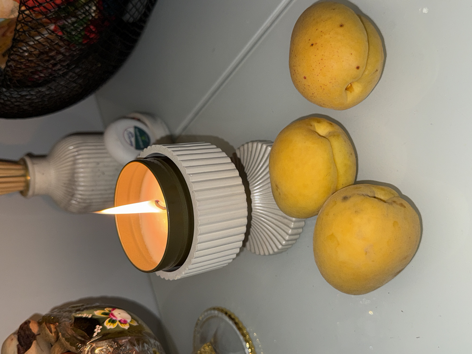An overhead, medium shot captures a lit candle in a cream-colored decorative holder, placed on a gray surface that appears to be a table. Three yellow fruits, possibly apricots, are arranged in front of the candle holder, two on the left and one on the right. In the background, a white ribbed vase with a wooden stick protruding from it stands to the left of the candle, and a black basket filled with colorful items is positioned to the right. To the far left, a glass bowl containing dried potpourri and decorative elements is partially visible. The lighting is dim, with the candle flame casting a warm glow on its surroundings.